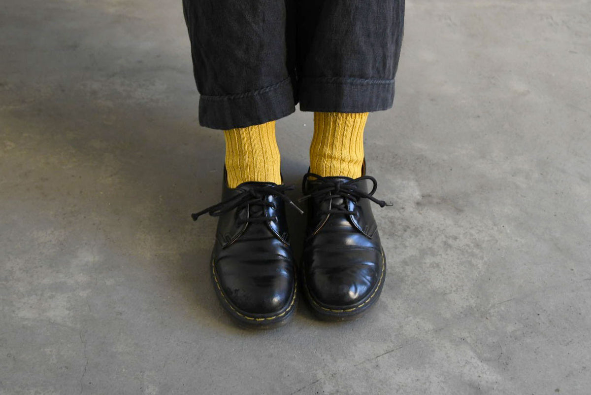 A person wearing black pants, Nishiguchi Kutsushita Praha Cotton Socks – Susuki-Gold, and black lace-up shoes stands on a gray concrete floor.