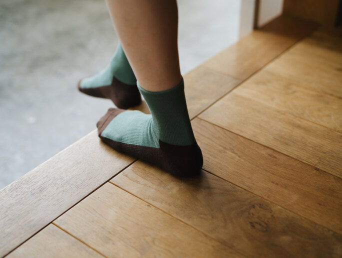 A person stands on tiptoe on a wooden floor near a step, wearing Nishiguchi Kutsushita Cotton Cashmere Walk Socks – Kusamochi: green women&#39;s socks with brown toes and heels.