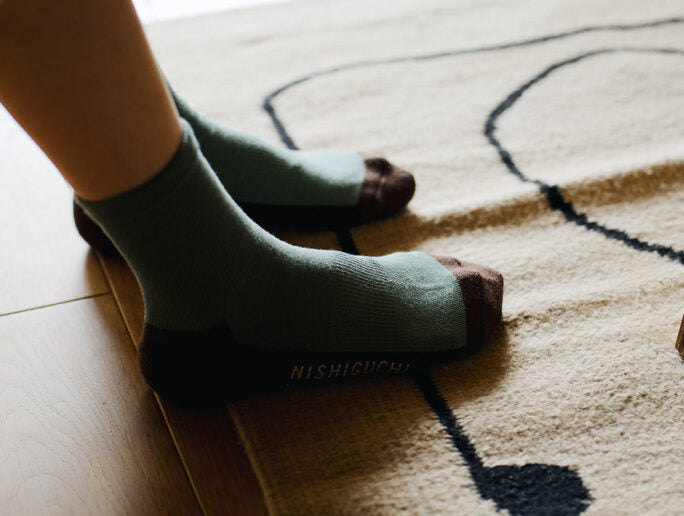 A person wearing Nishiguchi Kutsushita Cotton Cashmere Walk Socks – Kusamochi in green and brown stands on a beige rug with a dark blue abstract design—check our size chart for your perfect fit.