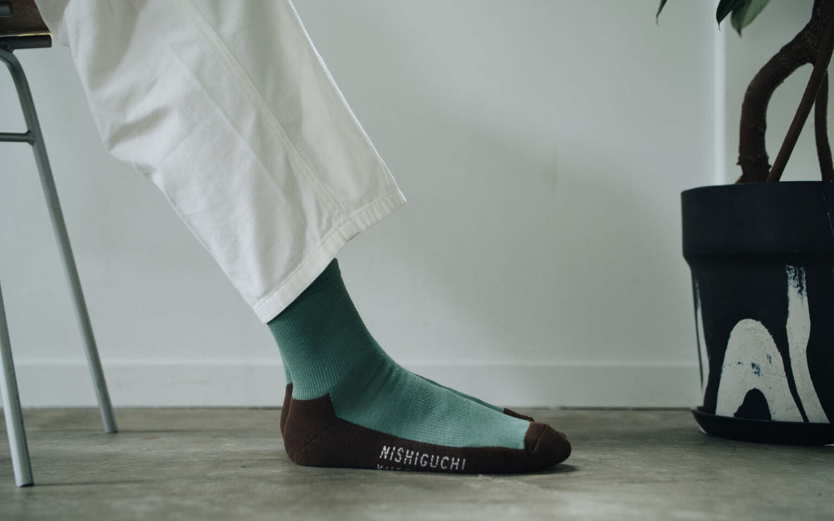 A person in white pants sits on a chair next to a black potted plant, wearing Nishiguchi Kutsushita Cotton Cashmere Walk Socks – Kusamochi in green and brown.