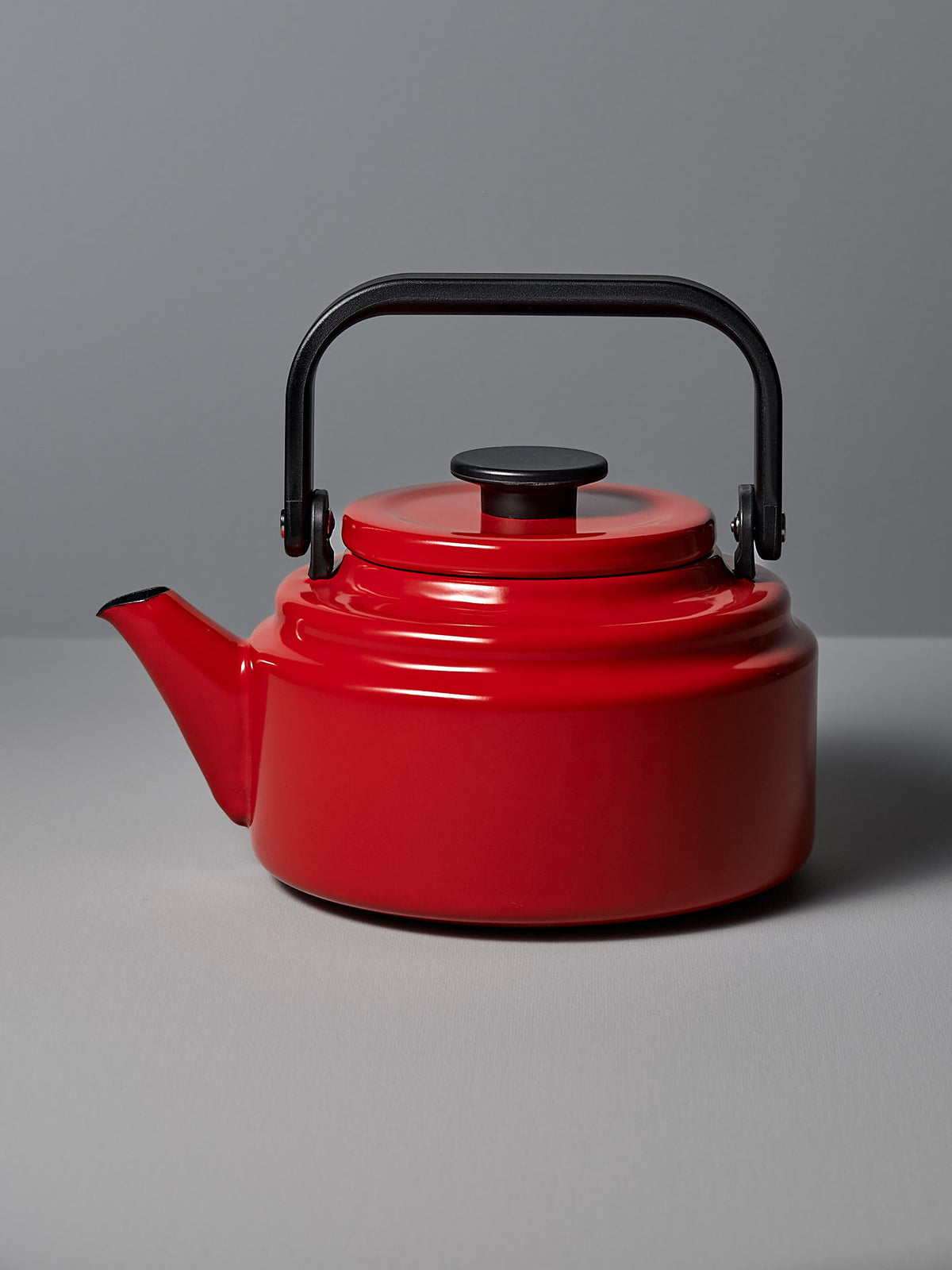 A red Amu Stove-top Kettle by Noda Horo, featuring a black handle and lid, rests on a gray surface against a gray backdrop, reflecting the elegance of traditional Japanese kitchens.