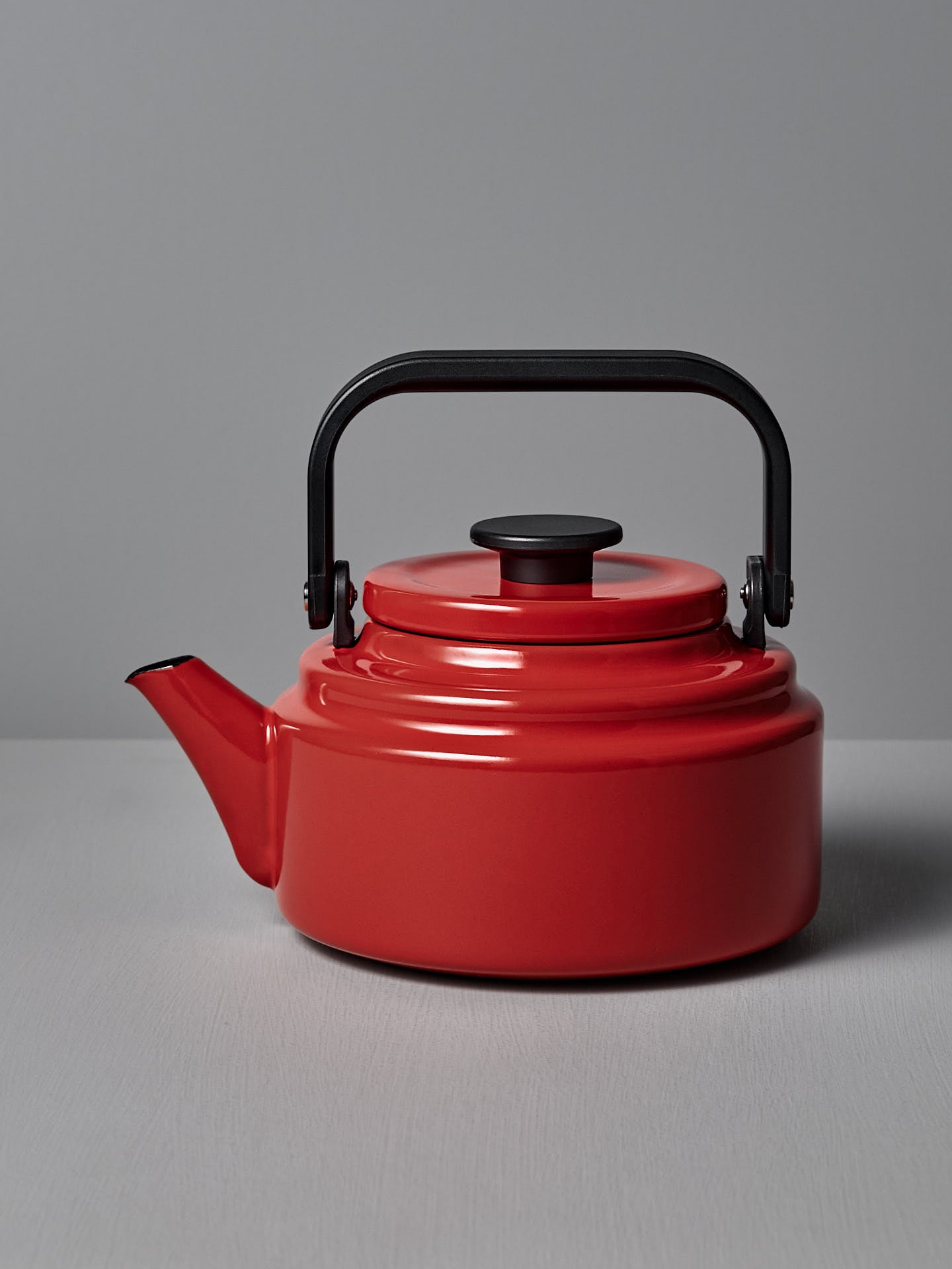 A red Amu Stove-top Kettle by Noda Horo, featuring a black handle and lid, rests on a gray surface against a gray backdrop, reflecting the elegance of traditional Japanese kitchens.