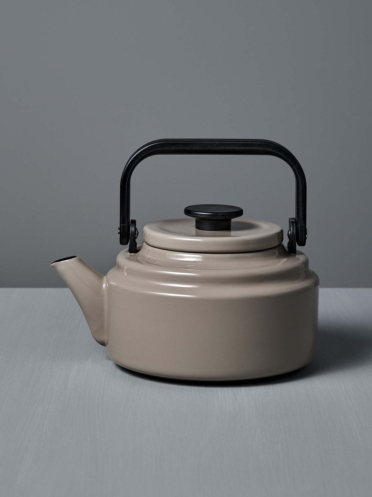 An Amu Stove-top Kettle – Beige by Noda Horo, featuring a black handle and lid, sits on a gray surface against a gray background.
