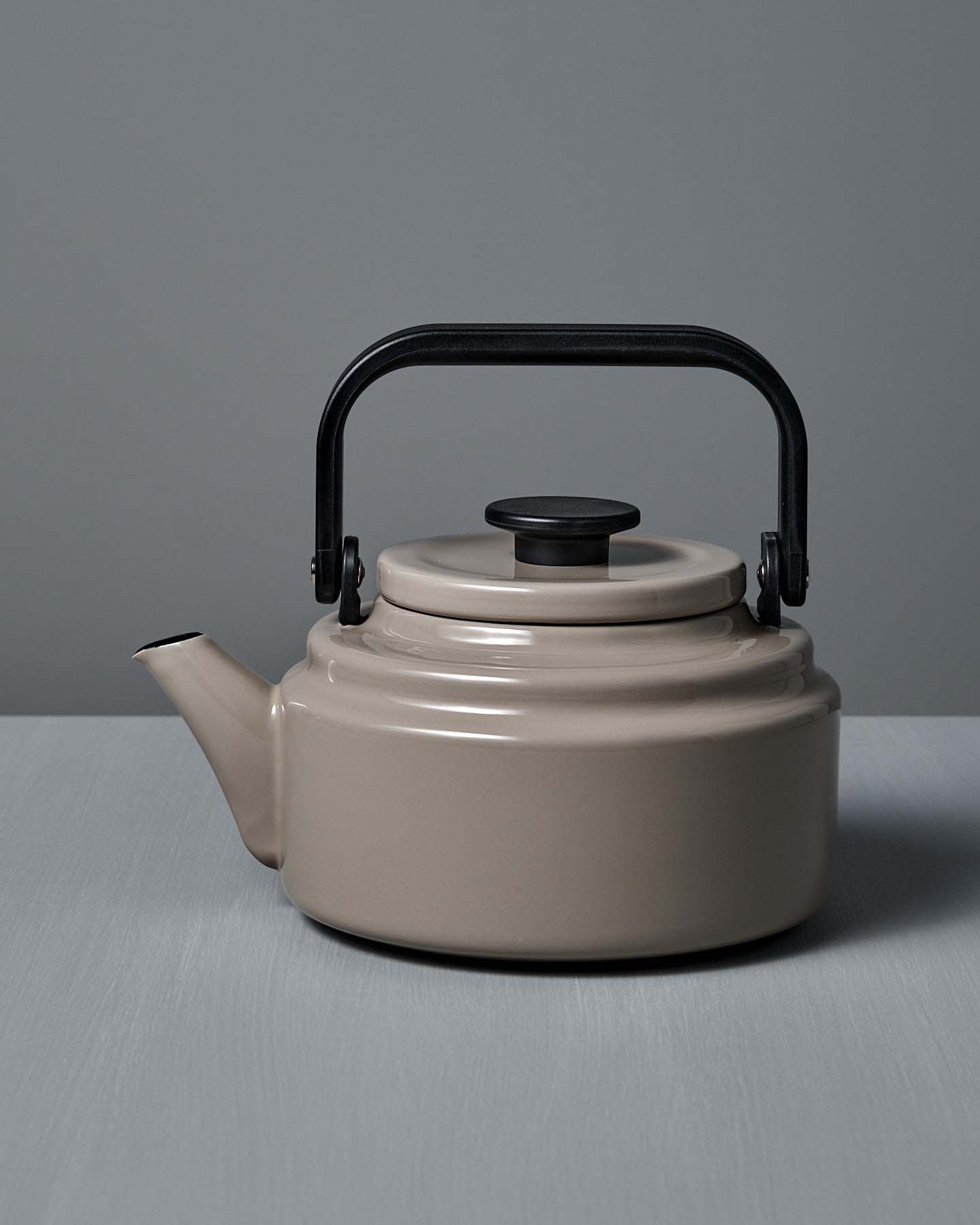 An Amu Stove-top Kettle – Beige by Noda Horo, featuring a black handle and lid, sits on a gray surface against a gray background.