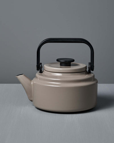 An Amu Stove-top Kettle – Beige by Noda Horo, featuring a black handle and lid, sits on a gray surface against a gray background.