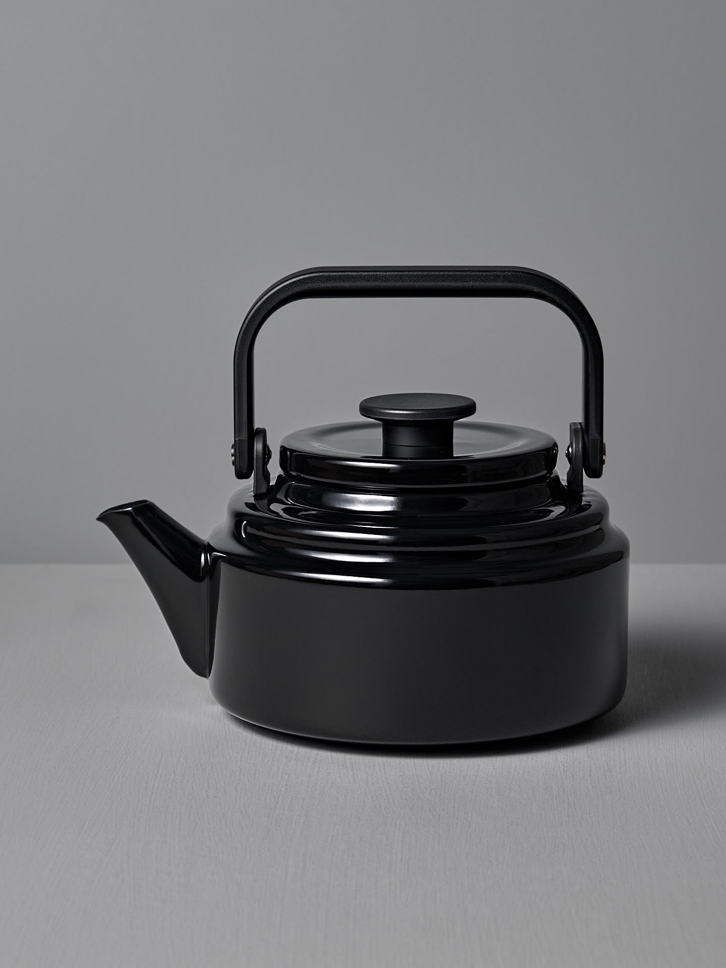 The Amu Stove-top Kettle – Black by Noda Horo, elegantly crafted from enamelware with a curved spout and rounded handle, is beautifully showcased on a gray surface against a matching gray background.
