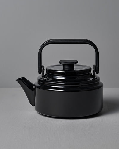 The Amu Stove-top Kettle – Black by Noda Horo, elegantly crafted from enamelware with a curved spout and rounded handle, is beautifully showcased on a gray surface against a matching gray background.