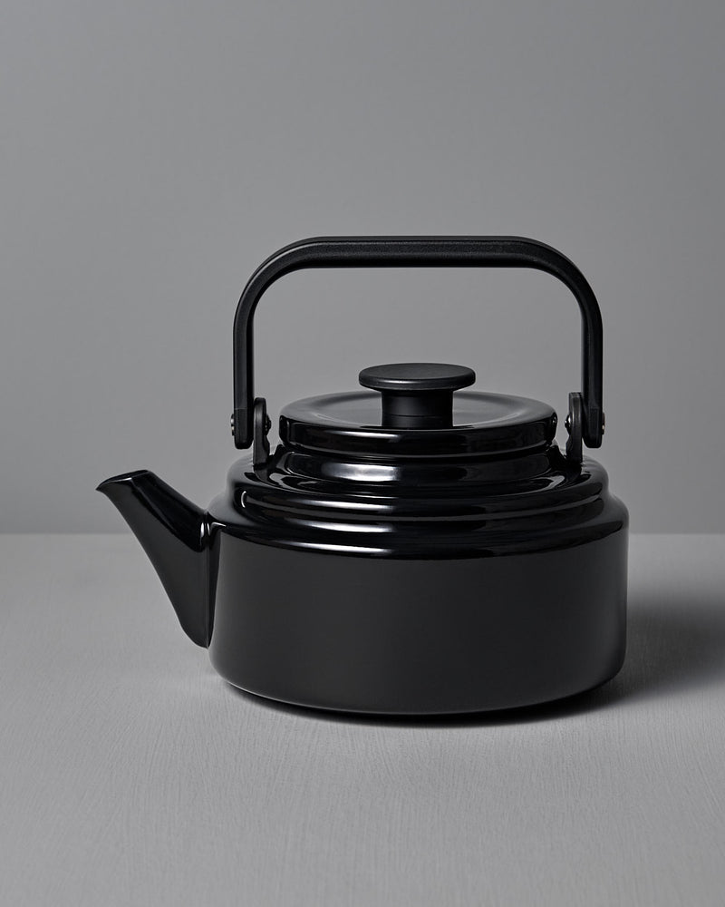 The Amu Stove-top Kettle – Black by Noda Horo, elegantly crafted from enamelware with a curved spout and rounded handle, is beautifully showcased on a gray surface against a matching gray background.