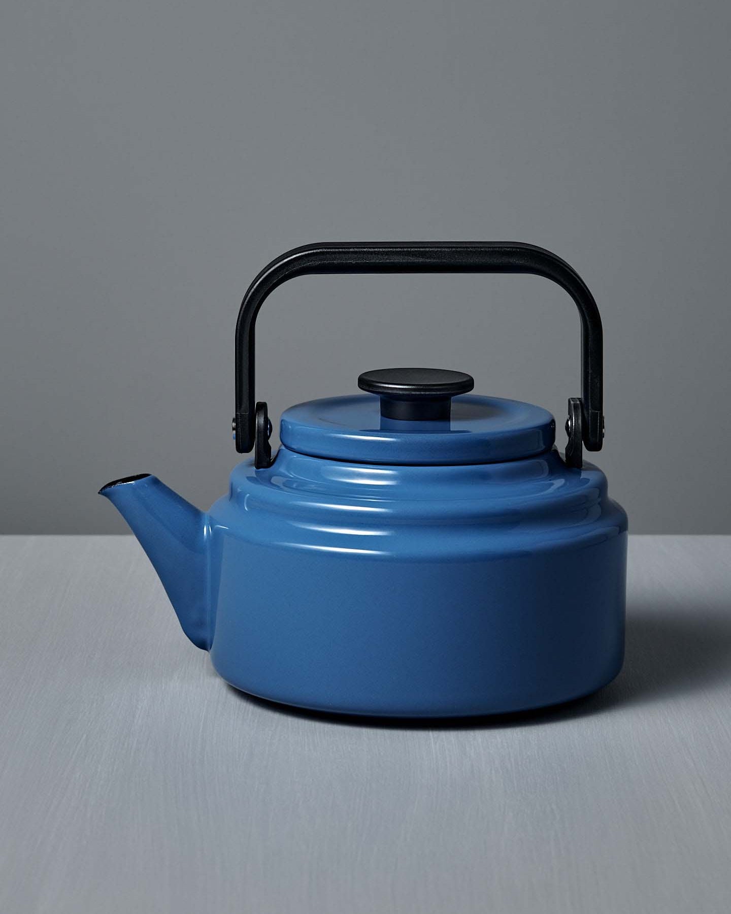 The Amu Stove-top Kettle – Blue by Noda Horo, featuring an enamelware finish and black handle and spout, sits on a gray surface against a gray background, evoking a classic Japanese design.