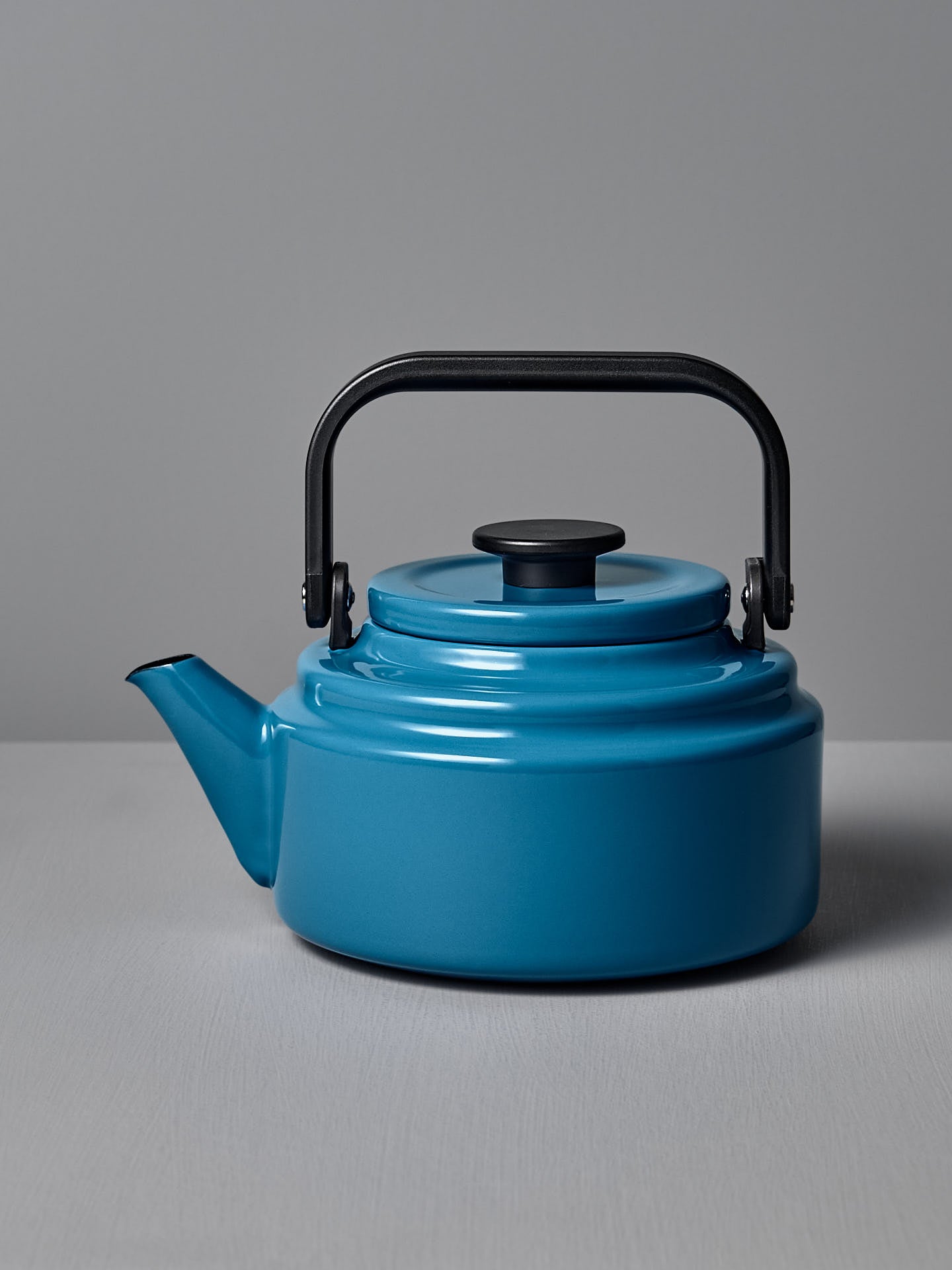The Amu Stove-top Kettle – Blue by Noda Horo, featuring an enamelware finish and black handle and spout, sits on a gray surface against a gray background, evoking a classic Japanese design.