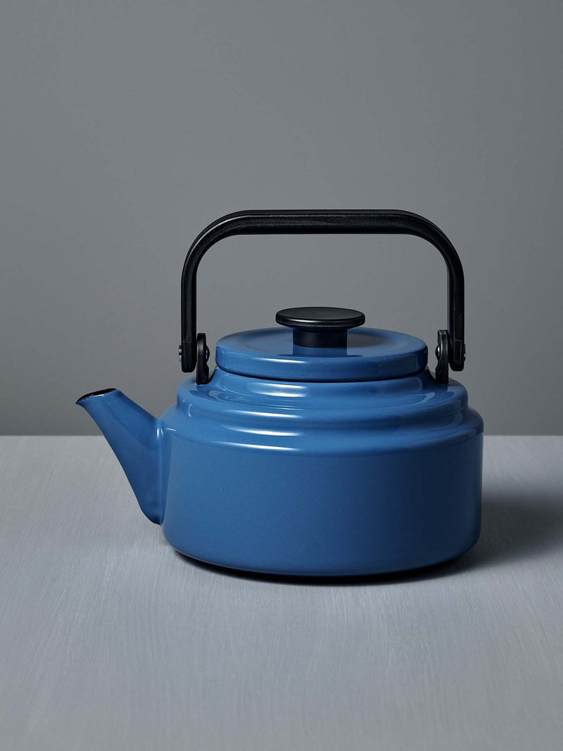 The Amu Stove-top Kettle – Blue by Noda Horo, featuring an enamelware finish and black handle and spout, sits on a gray surface against a gray background, evoking a classic Japanese design.