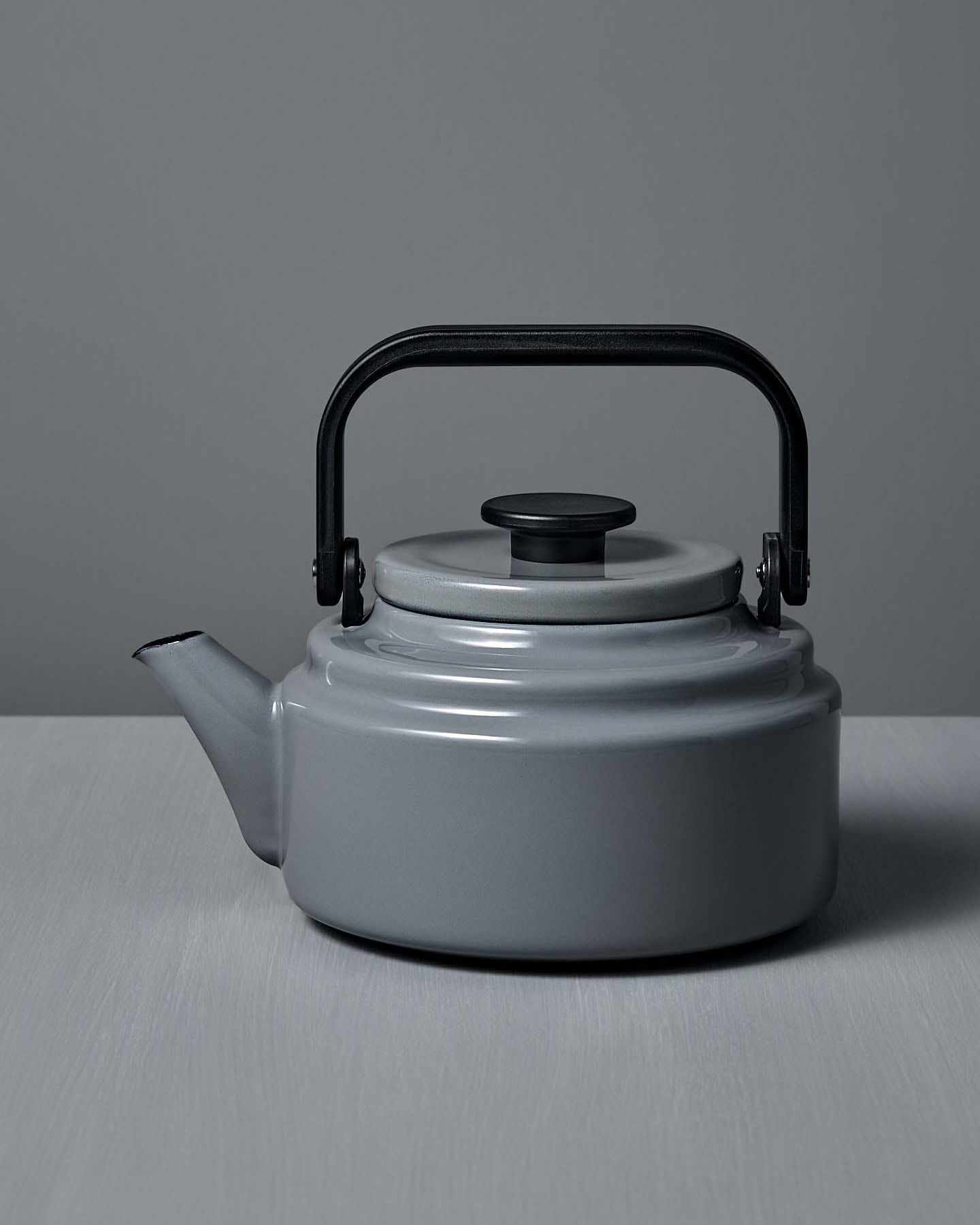 The Amu Stove-top Kettle – Grey, with its black handle, rests on a light gray surface against a matching background, highlighting the refined design of Japanese enamelware.