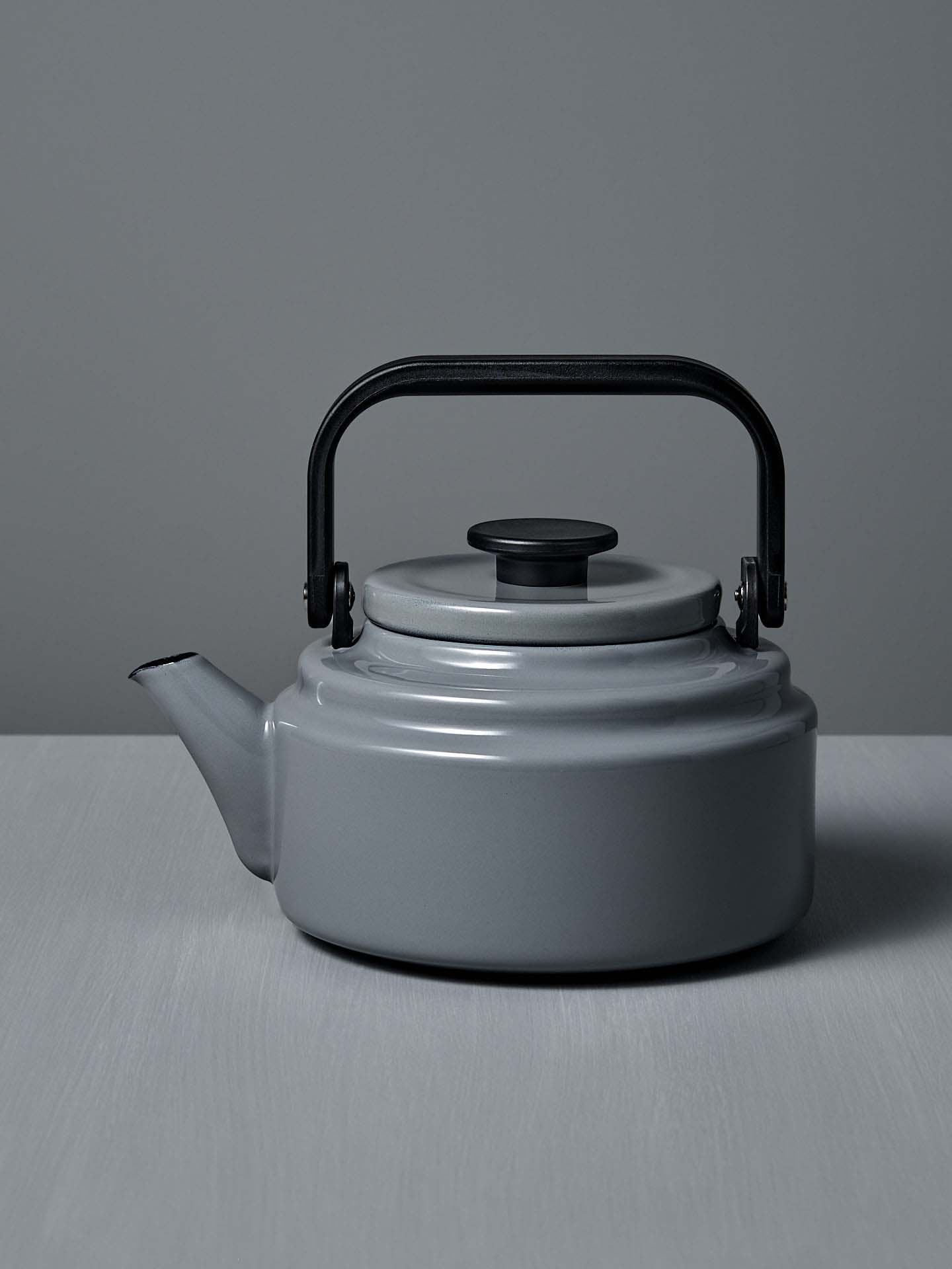 The Amu Stove-top Kettle – Grey, with its black handle, rests on a light gray surface against a matching background, highlighting the refined design of Japanese enamelware.