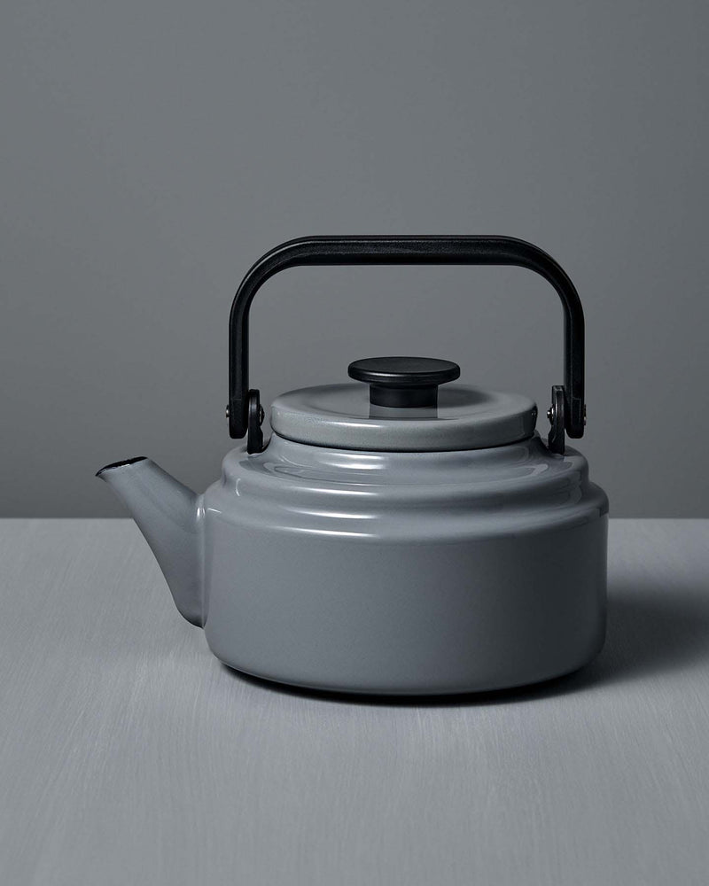 The Amu Stove-top Kettle – Grey, with its black handle, rests on a light gray surface against a matching background, highlighting the refined design of Japanese enamelware.