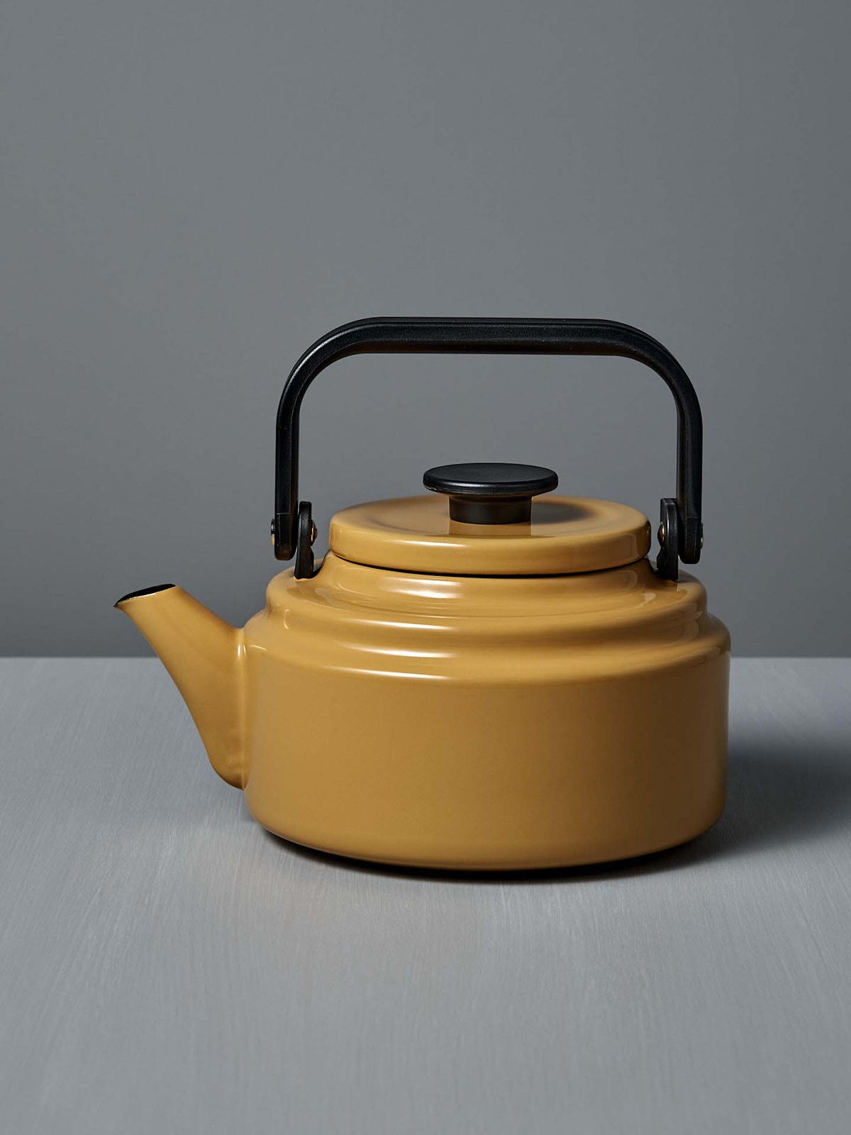 The Amu Stove-top Kettle – Yellow, a vibrant Japanese enamel kettle with a black handle and lid, sits on a light gray surface against a gray background.