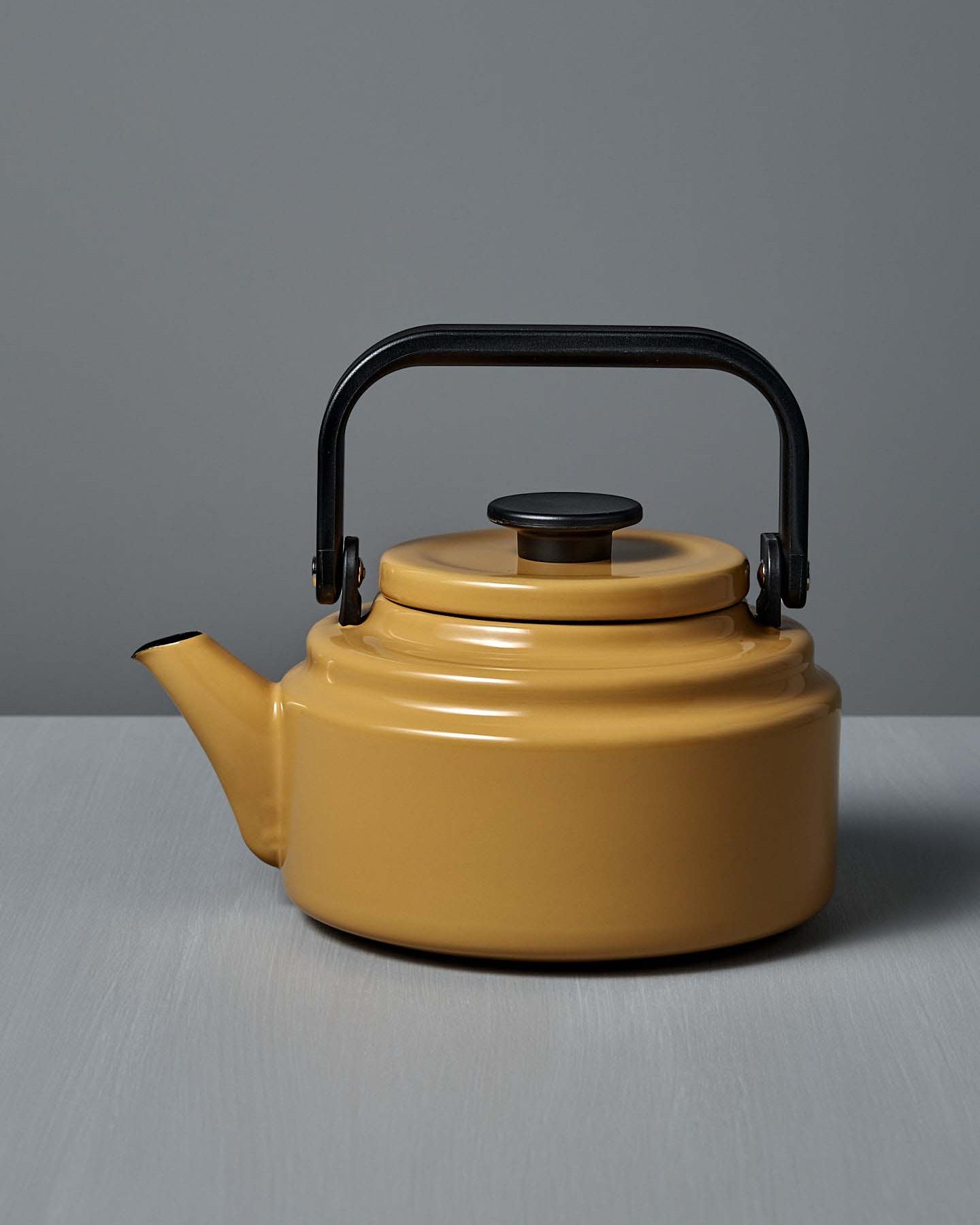 The Amu Stove-top Kettle – Yellow, a vibrant Japanese enamel kettle with a black handle and lid, sits on a light gray surface against a gray background.