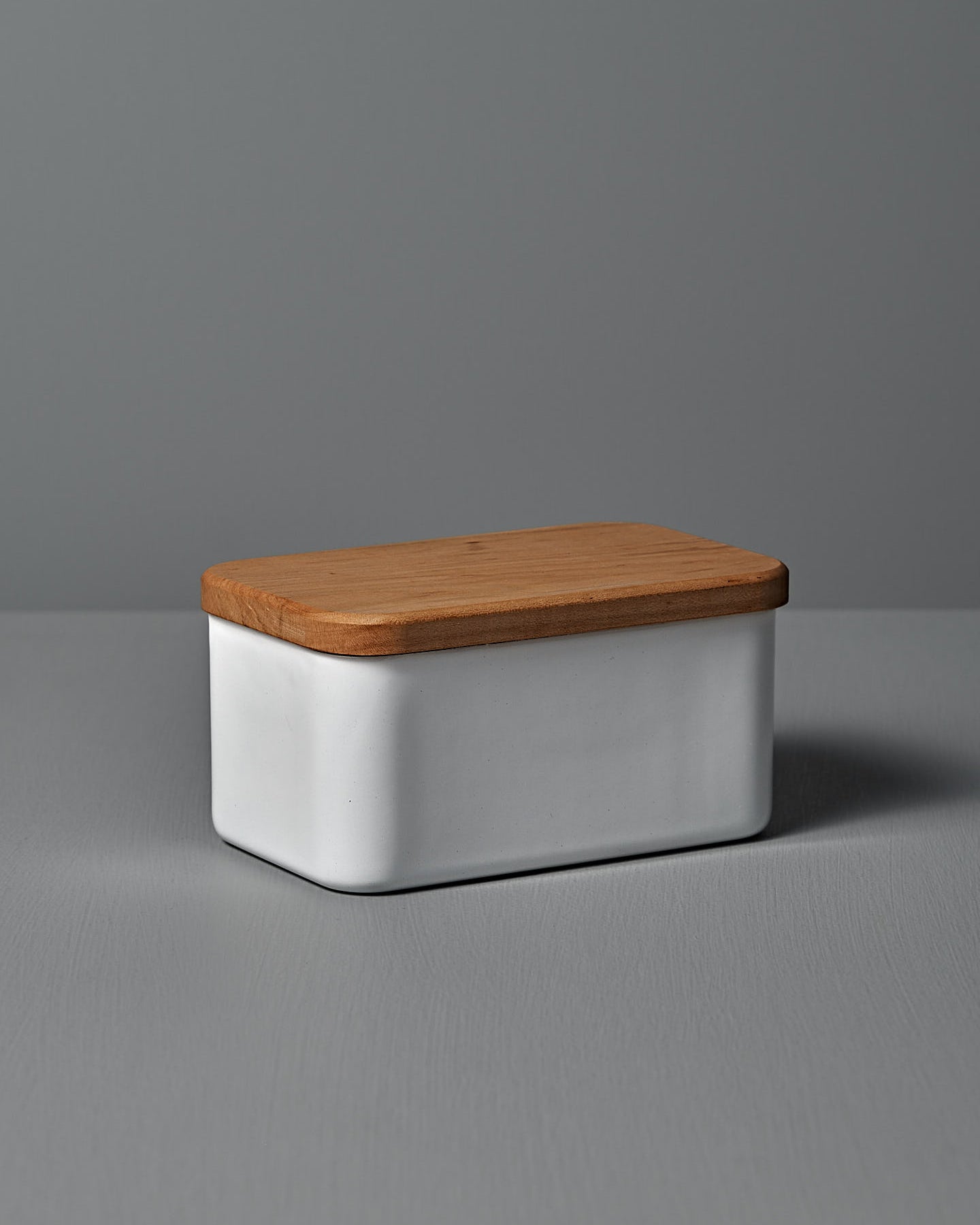 The Enamel Butter Case – 450g by Noda Horo, a rectangular white ceramic container with a Japanese cherry wood lid, rests elegantly on a gray surface against a soft gray backdrop.