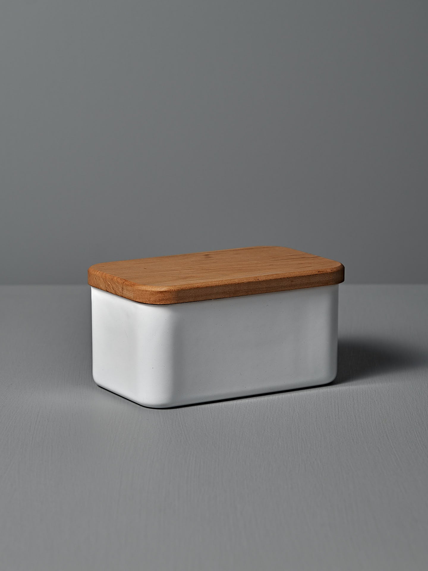 The Enamel Butter Case – 450g by Noda Horo, a rectangular white ceramic container with a Japanese cherry wood lid, rests elegantly on a gray surface against a soft gray backdrop.