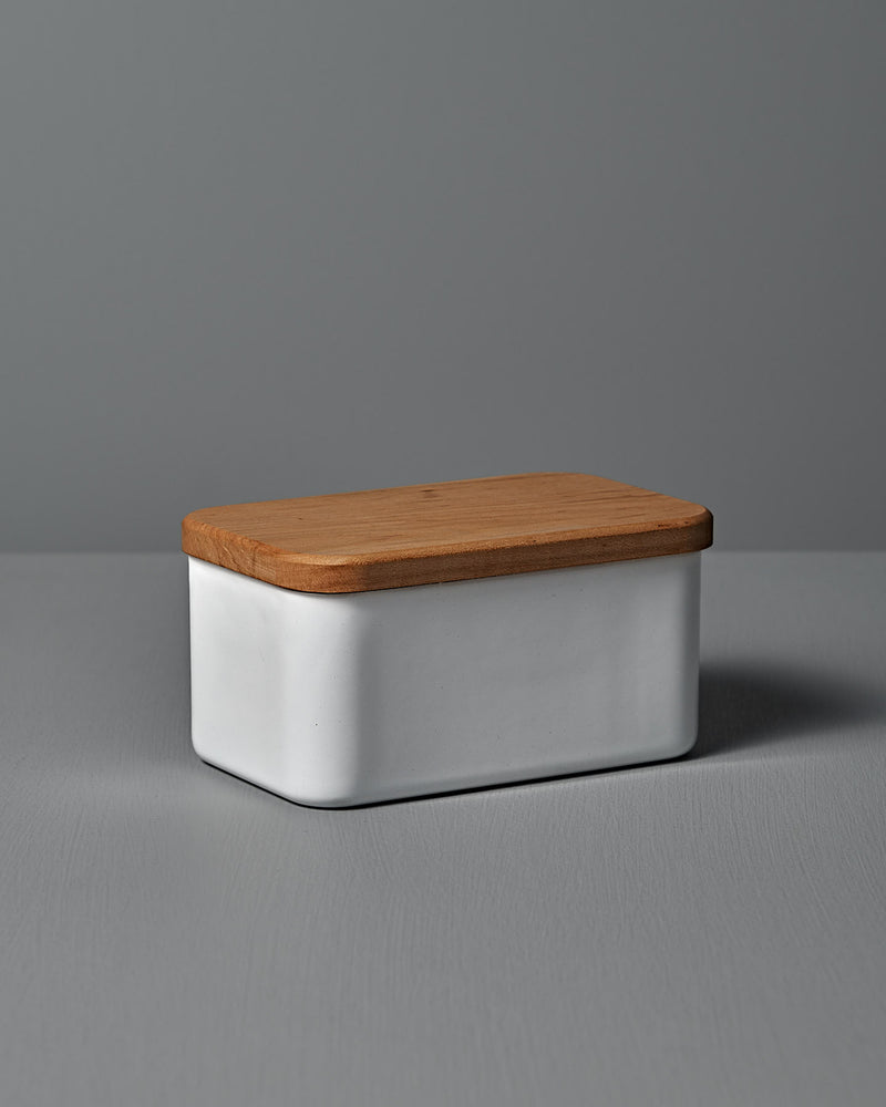 The Enamel Butter Case – 450g by Noda Horo, a rectangular white ceramic container with a Japanese cherry wood lid, rests elegantly on a gray surface against a soft gray backdrop.