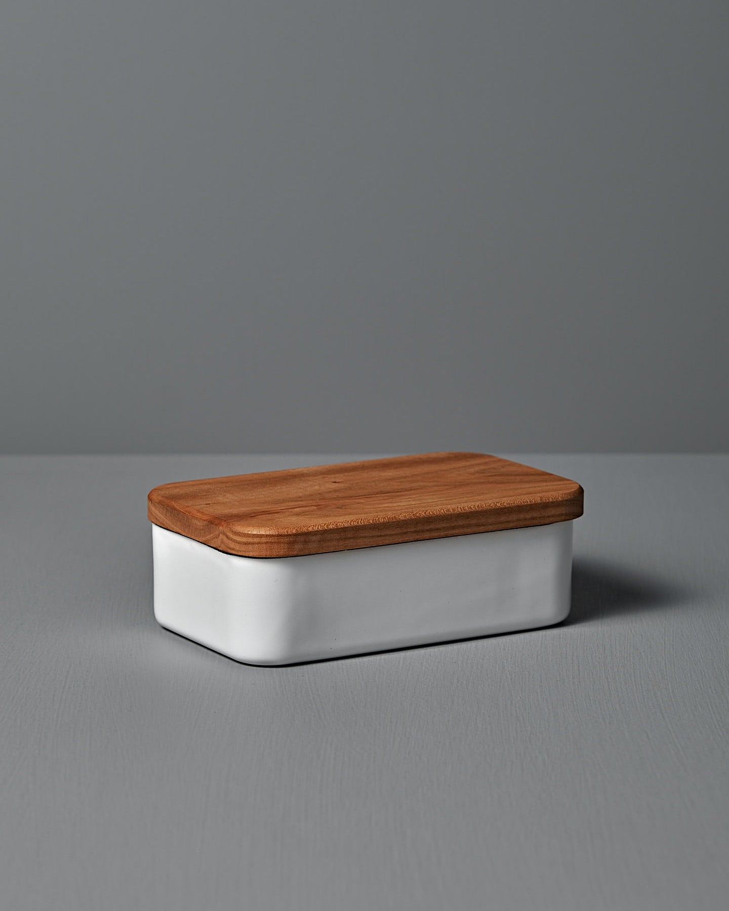 The Noda Horo Enamel Butter Case – 200g, with its sleek white ceramic body and cherry wood lid, rests elegantly on a gray surface, providing stylish food storage.