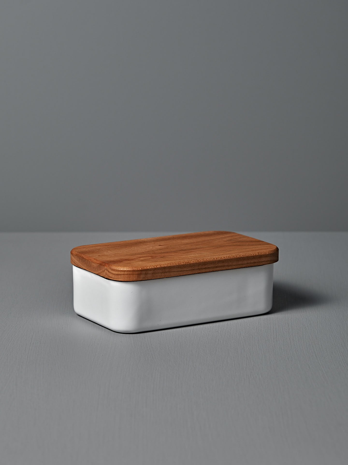 The Noda Horo Enamel Butter Case – 200g, with its sleek white ceramic body and cherry wood lid, rests elegantly on a gray surface, providing stylish food storage.