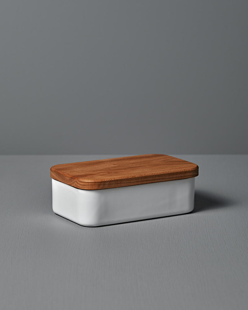 The Noda Horo Enamel Butter Case – 200g, with its sleek white ceramic body and cherry wood lid, rests elegantly on a gray surface, providing stylish food storage.