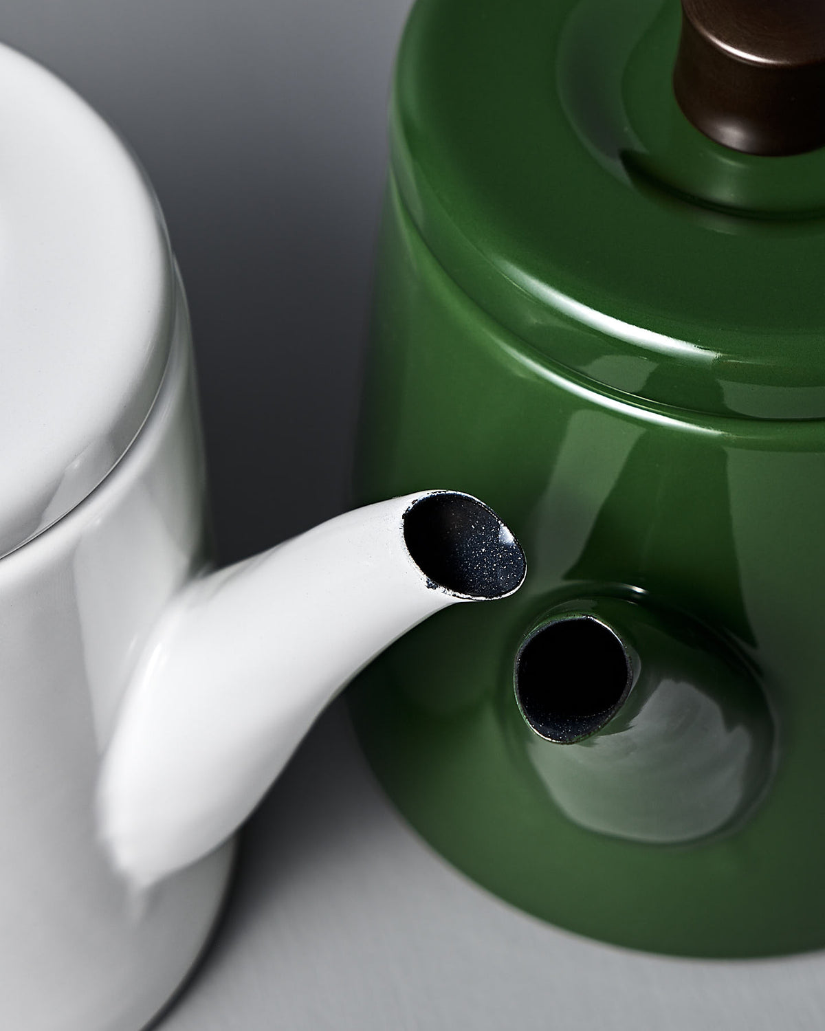A Potoru Stove-top Kettle/Teapot in green by Noda Horo paired with a white one, both have spouts pointing towards each other on a gray surface, highlighting the elegance of Japanese kitchenware.