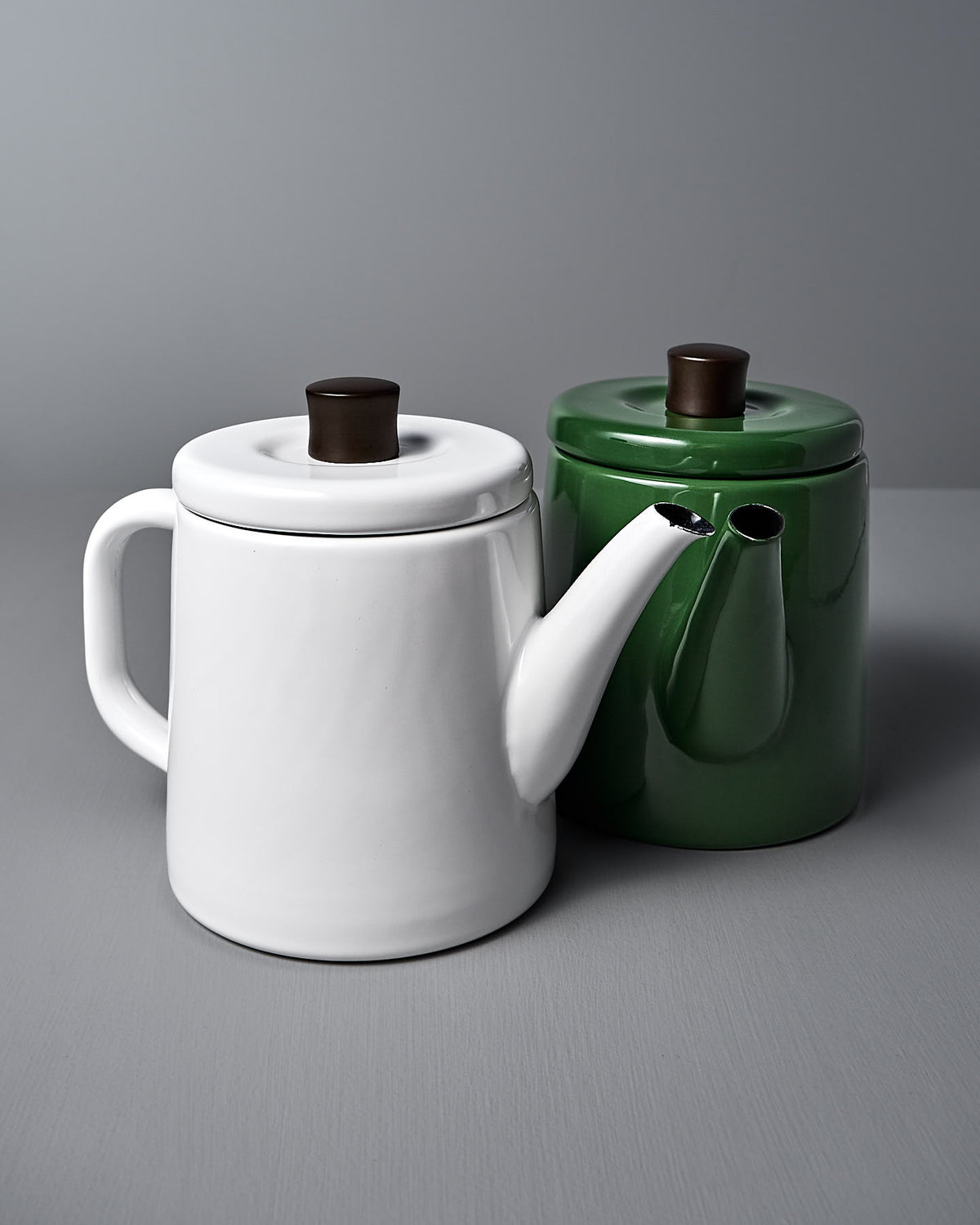 A Potoru Stove-top Kettle/Teapot by Noda Horo sits on a gray surface, white with a brown lid and spout, exuding traditional Japanese design. Beside it, a green teapot complements its modern elegance with simplicity.