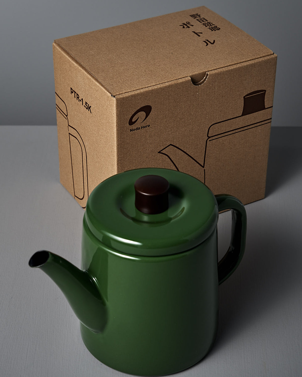A Noda Horo Potoru stove-top kettle/teapot in green with a dark brown lid is placed in front of a cardboard box featuring an illustration of the teapot, highlighting the elegance of Japanese kitchenware.