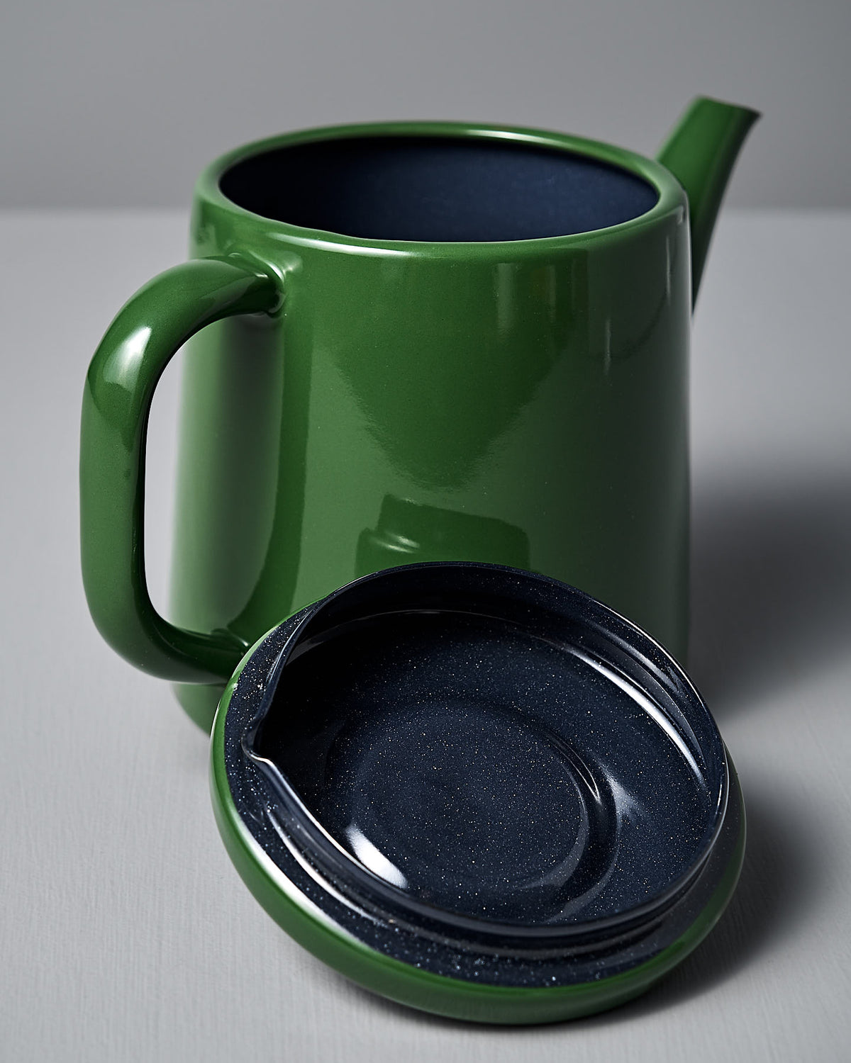 A Potoru Stove-top Kettle/Teapot by Noda Horo, in green enamelware with a wide handle and spout, sits on a light surface. Its dark blue lid is removed and placed in front of the kettle, highlighting the allure of Japanese kitchenware.