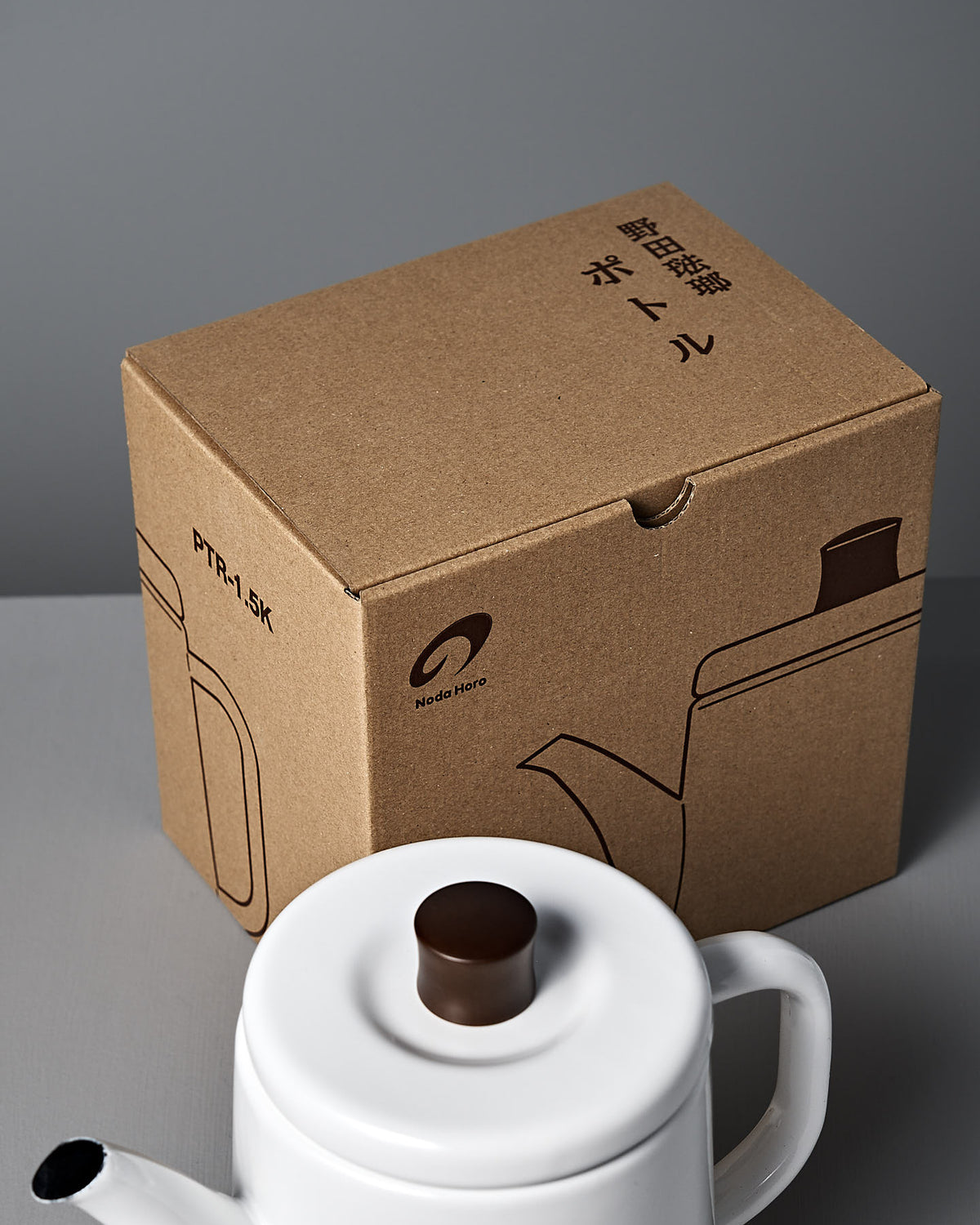 A Potoru Stove-top Kettle/Teapot – White by Noda Horo, featuring a dark brown lid, sits on a table beside a cardboard box with matching teapot illustration and Japanese text, epitomizing elegant Japanese kitchenware.
