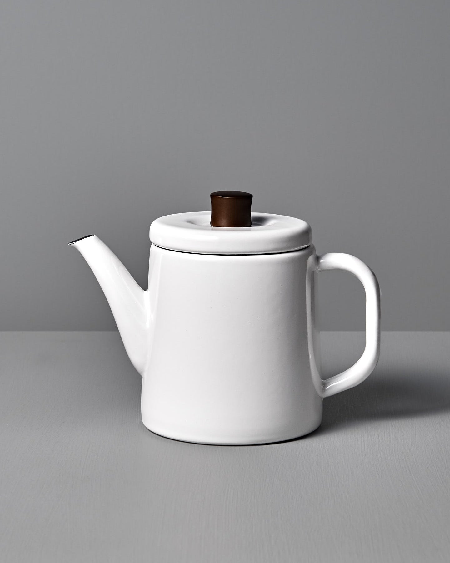 The Potoru Stove-top Kettle/Teapot – White, by Noda Horo, features a dark brown lid handle and is set elegantly on a gray surface against a gray backdrop, reminiscent of Japanese kitchenware.
