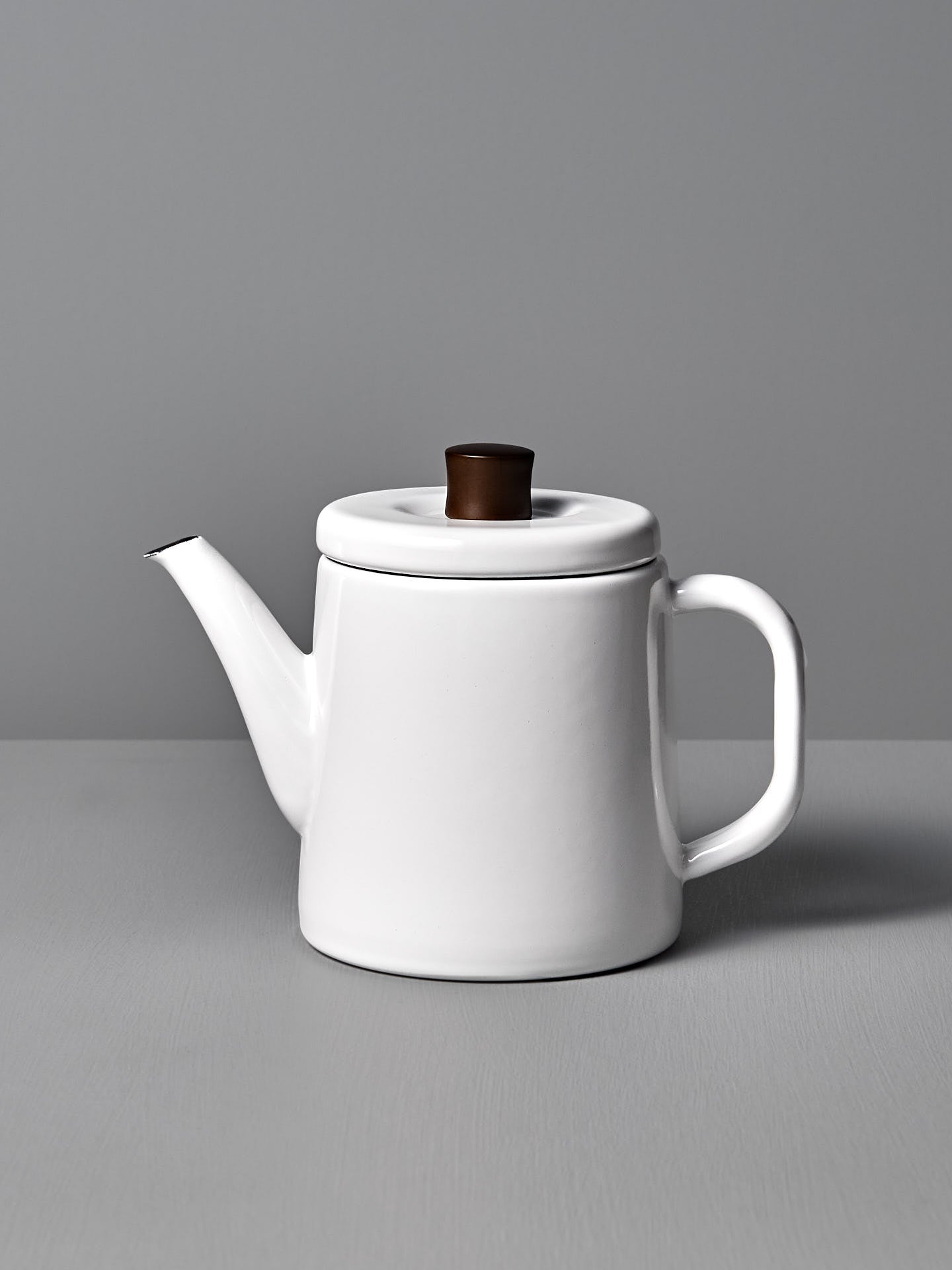 The Potoru Stove-top Kettle/Teapot – White, by Noda Horo, features a dark brown lid handle and is set elegantly on a gray surface against a gray backdrop, reminiscent of Japanese kitchenware.