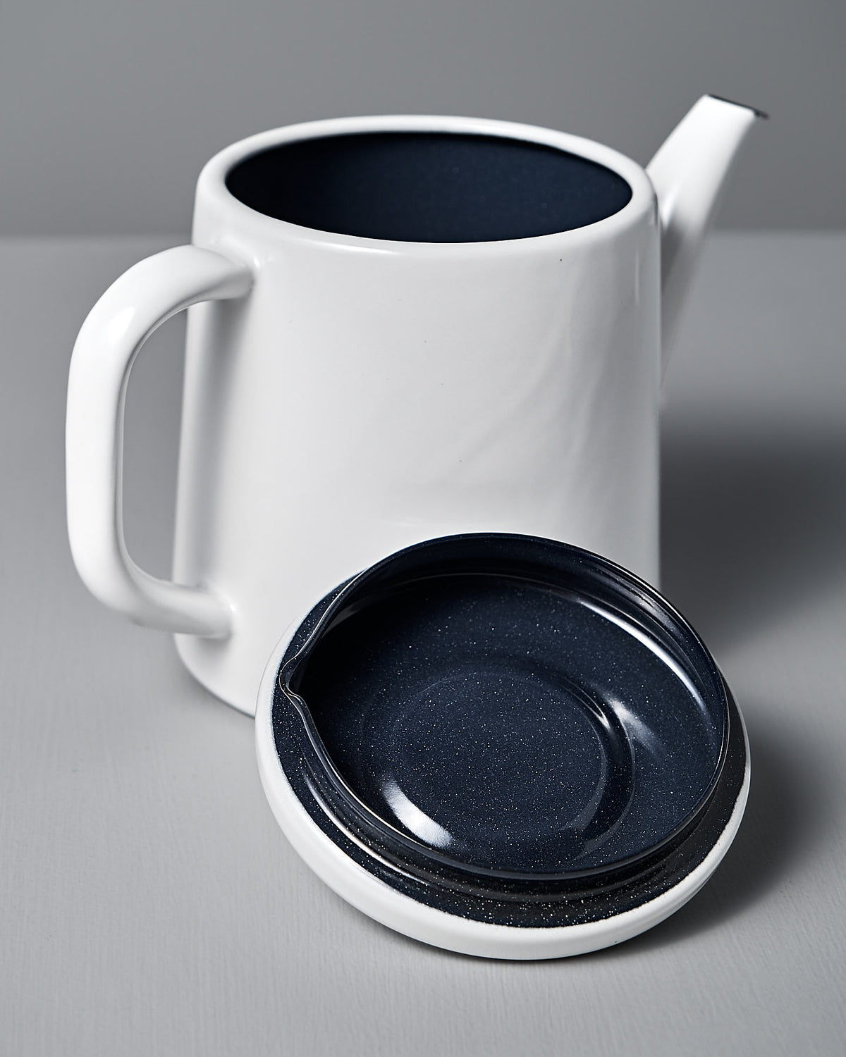 The Noda Horo Potoru Stove-top Kettle/Teapot in white sports a sleek handle and spout and a dark blue interior, with its matching lid beside it on a gray surface, showcasing the minimalist charm of Japanese kitchenware.