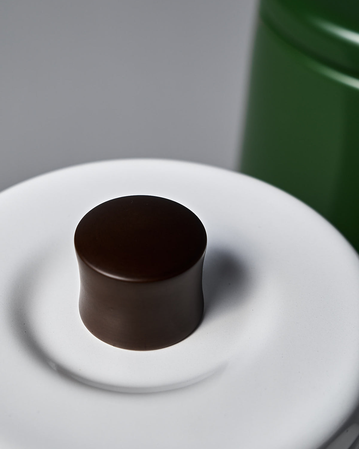A close-up of the Potoru Stove-top Kettle/Teapot – White by Noda Horo shows its white ceramic lid with a dark brown knob, alongside a green cylindrical object, evoking Japanese kitchenware.