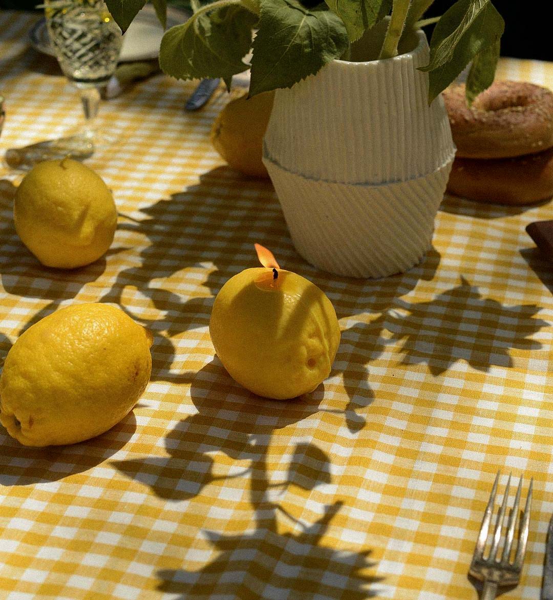 A Scented Lemon Candle from Nonna&#39;s Grocer illuminates a yellow checkered tablecloth with a white vase and bagels, as plant shadows playfully dance, creating an ambiance of elegance akin to handmade candles.