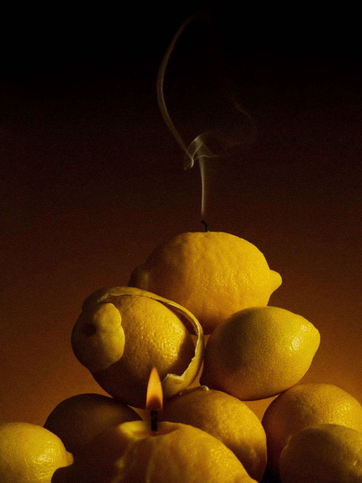 A stack of lemons in dim lighting features Nonna&#39;s Grocer Scented Lemon Candle at the top emitting smoke and another at the bottom with a flickering flame.