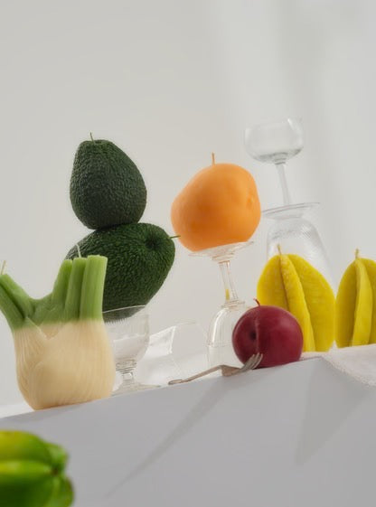 A Nonna&#39;s Grocer handmade mango candle crafted from a soy wax blend sits amidst a group of fruits and vegetables on a table.