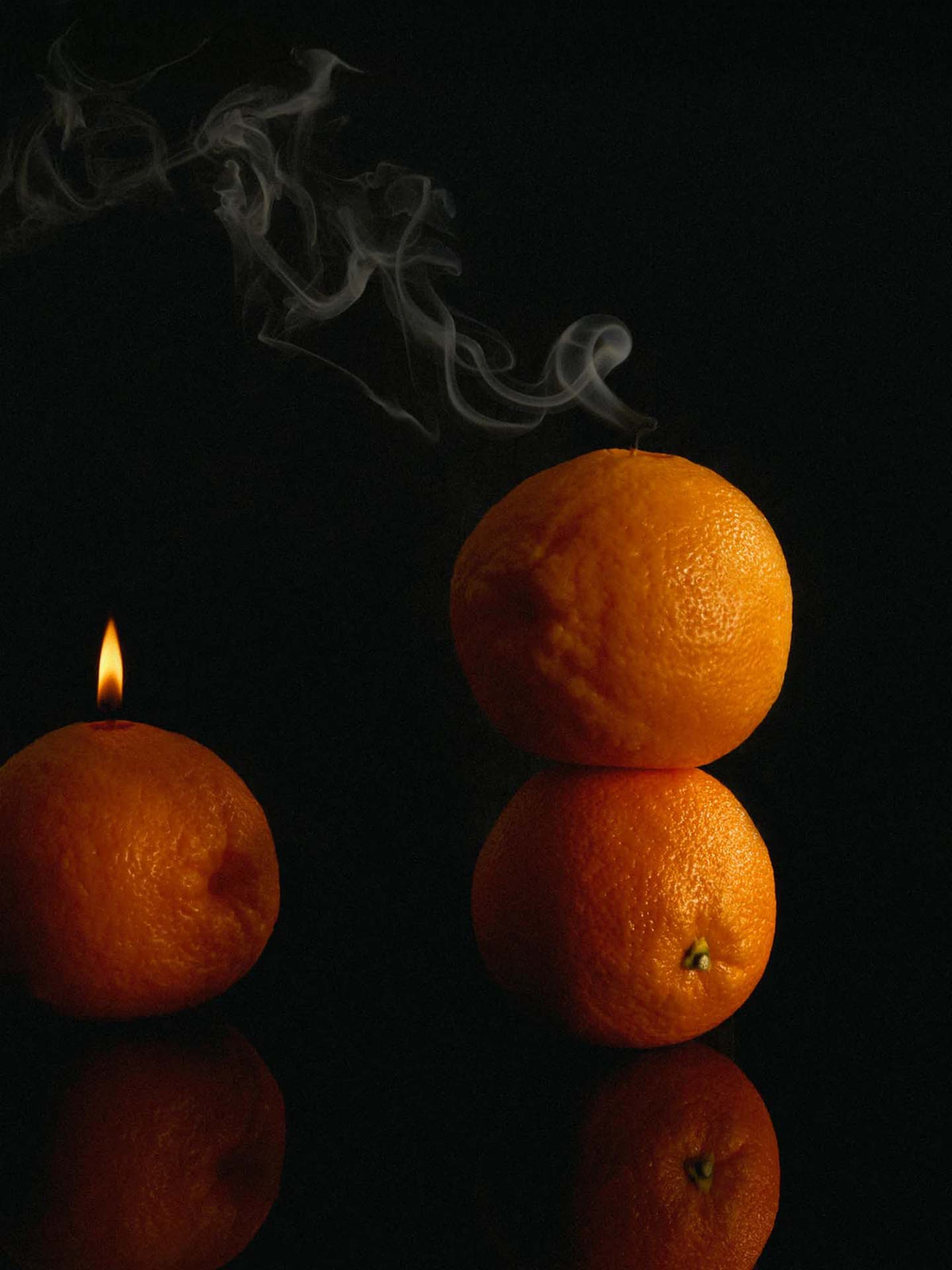 A stack of two oranges rests beside an orange adorned with Nonna&#39;s Grocer&#39;s Scented Orange Candle, its flame casting a warm glow against the dark backdrop. Wisps of rising smoke add to the atmosphere, highlighting notes of natural soy wax and orange peel essence.