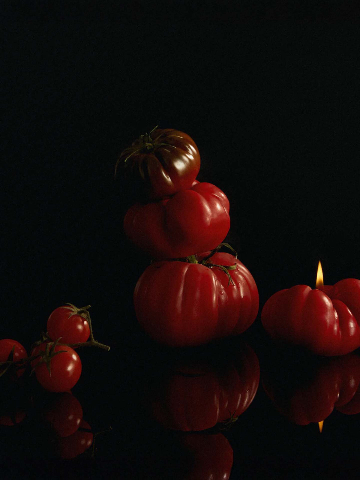 A stack of red tomatoes in different sizes is arranged on a black reflective surface, highlighting the warm glow of Nonna&#39;s Grocer Scented Tomato Candle&#39;s gentle flame on the right.