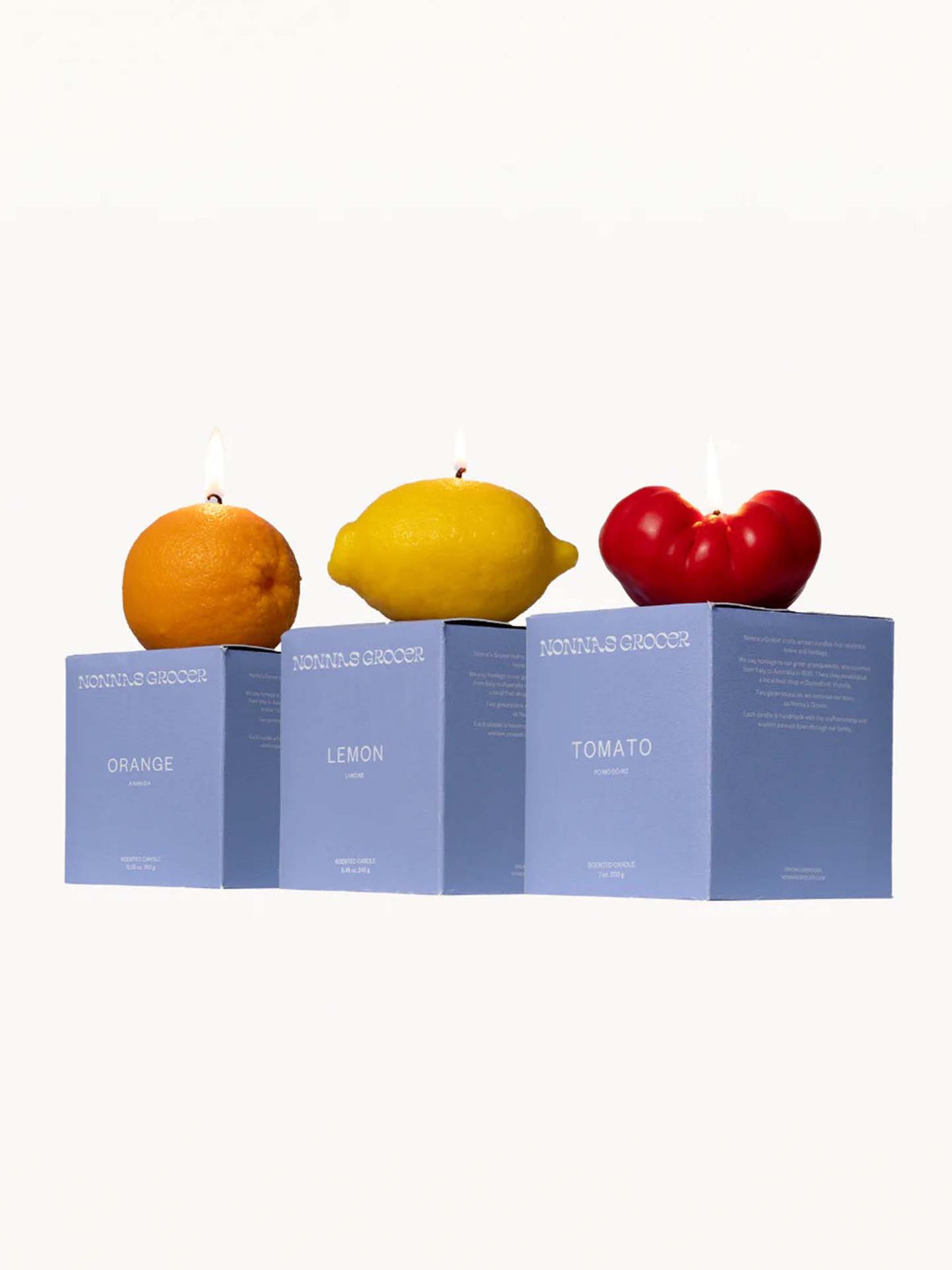 The Scented Trio Pack from Nonna&#39;s Grocer features candles shaped like an orange, lemon, and tomato, each beautifully lit atop matching blue boxes labeled &quot;Orange,&quot; &quot;Lemon,&quot; and &quot;Tomato.&quot; Crafted from natural soy wax, these delightful creations are perfect surprises for gift boxes.