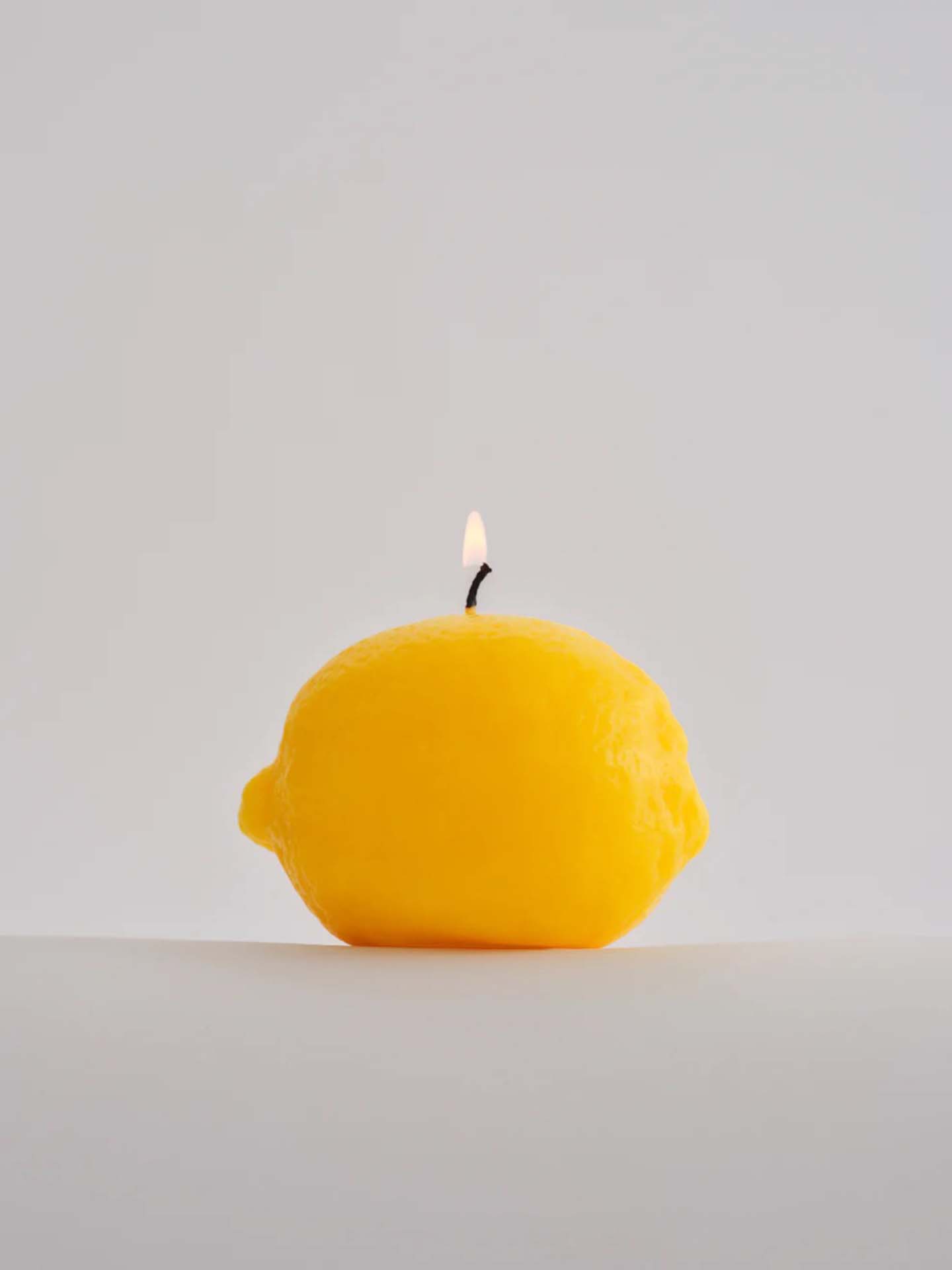 A Scented Lemon Candle from Nonna's Grocer, featuring a handmade design in the shape of a lemon, made from natural soy wax with a lit wick set against a plain background.