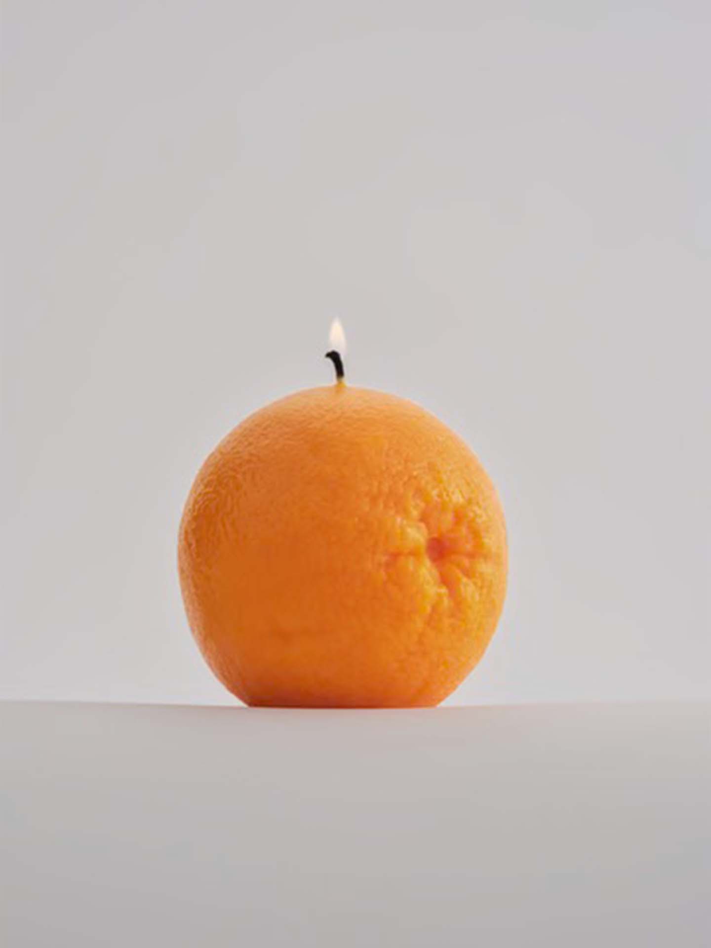 A Scented Orange Candle by Nonna's Grocer, featuring a small lit wick, set against a plain background.