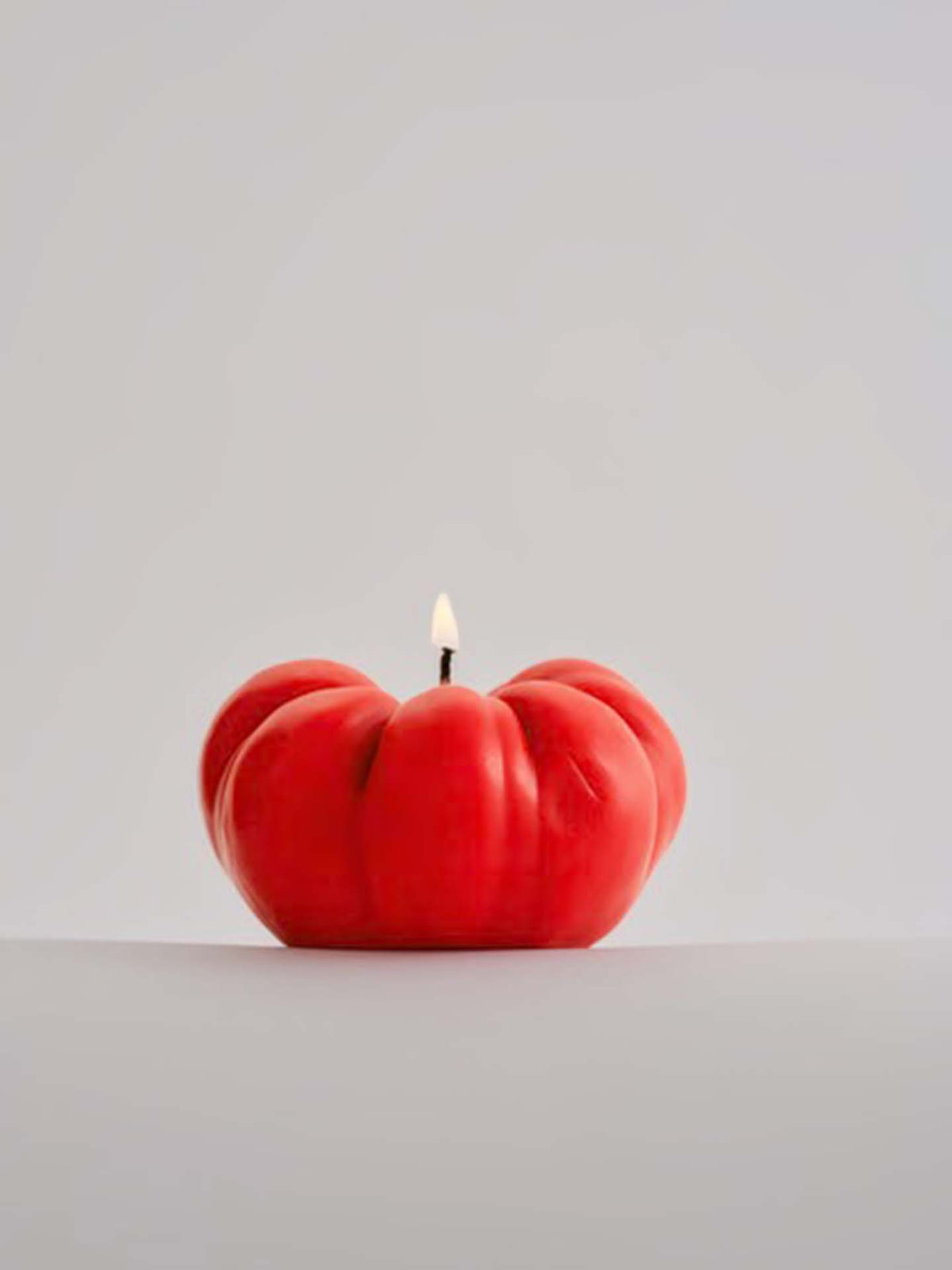 A red, tomato-shaped candle from Nonna&#39;s Grocer made from natural soy wax with a lit flame against a plain gray background.