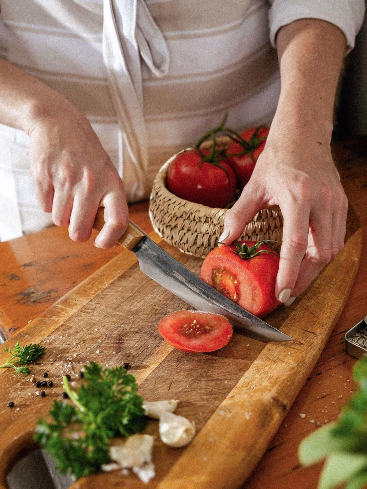 A person slices a tomato on a wooden board with the №2 – Farmer Knife; parsley, garlic, and peppercorns are also on the board. A basket of tomatoes sits in the background.