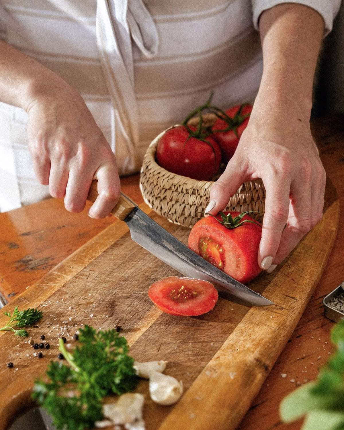 A person slices a tomato on a wooden board with the №2 – Farmer Knife; parsley, garlic, and peppercorns are also on the board. A basket of tomatoes sits in the background.
