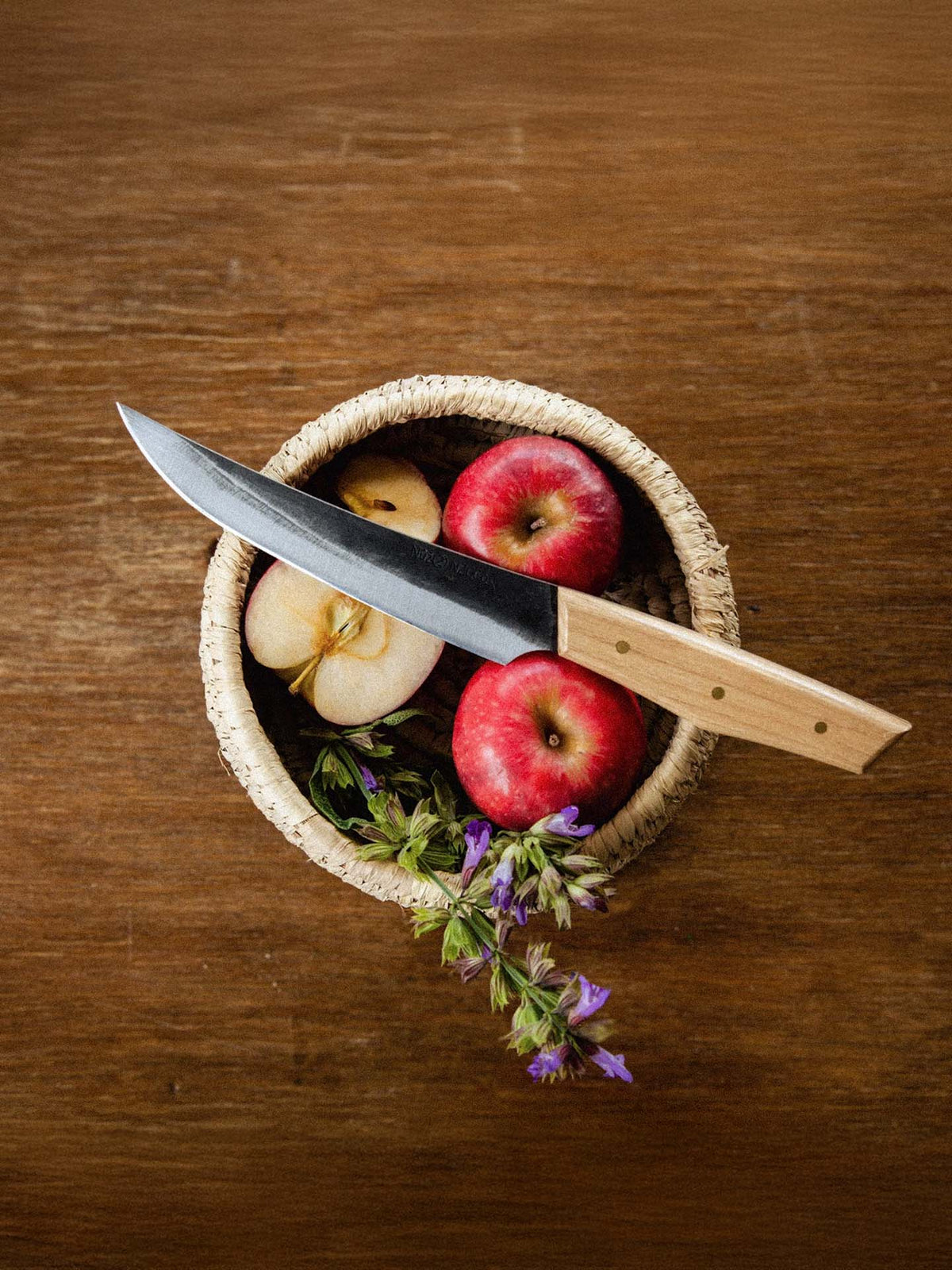 On a wooden surface sits a woven basket with two whole apples, a halved apple, purple flowers, and the №2 – Farmer Knife featuring Swedish carbon steel and an American Ash handle.
