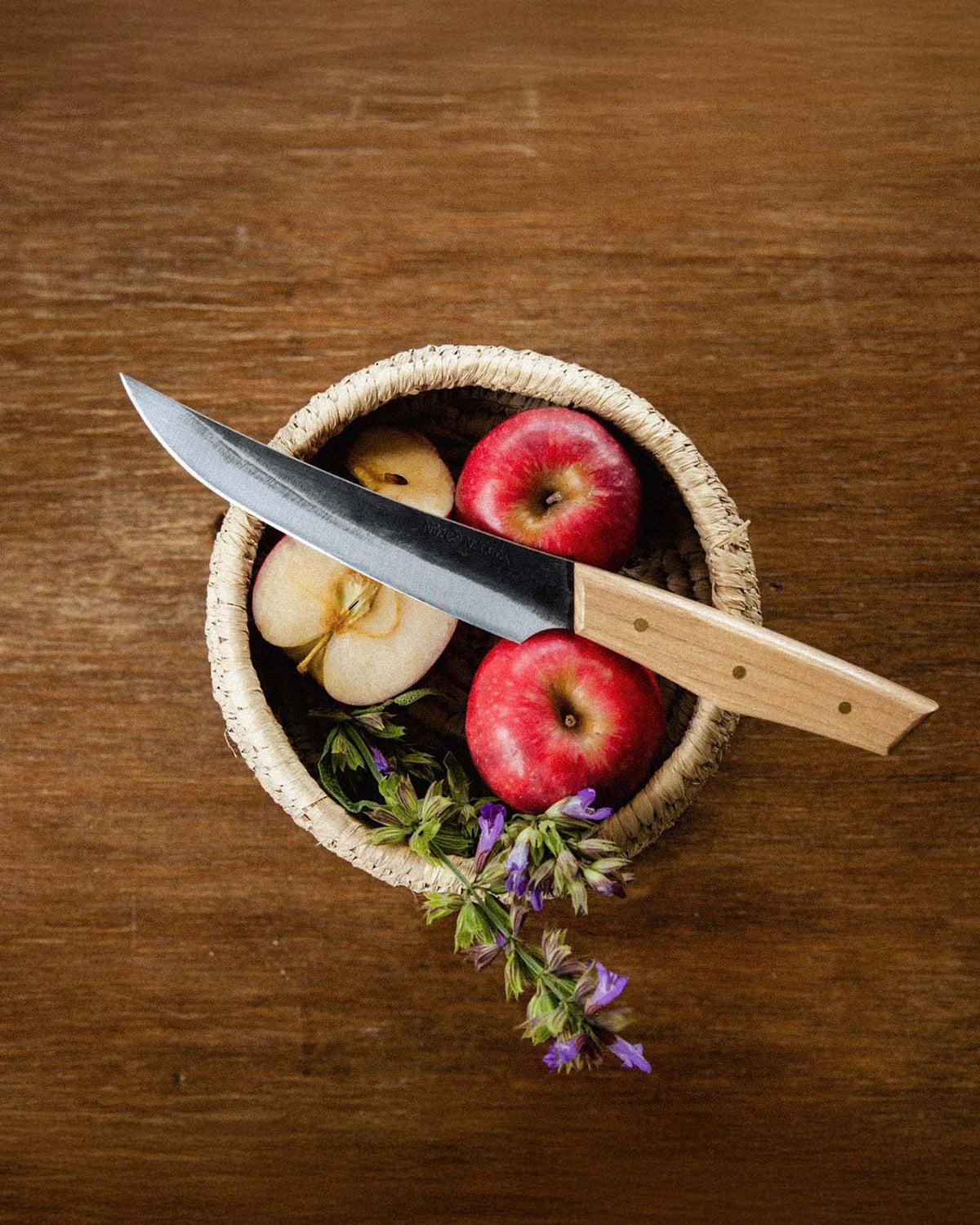 On a wooden surface sits a woven basket with two whole apples, a halved apple, purple flowers, and the №2 – Farmer Knife featuring Swedish carbon steel and an American Ash handle.