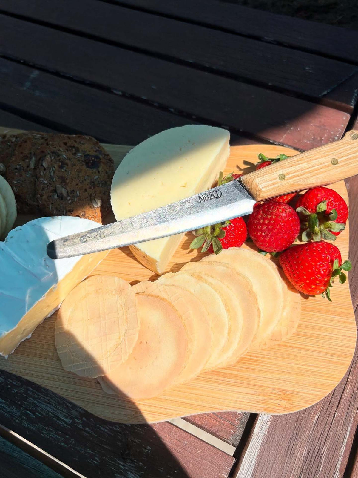 The №1 – Spread Knife rests on a wooden board with assorted cheeses, round crackers, and fresh strawberries, set atop an outdoor table.