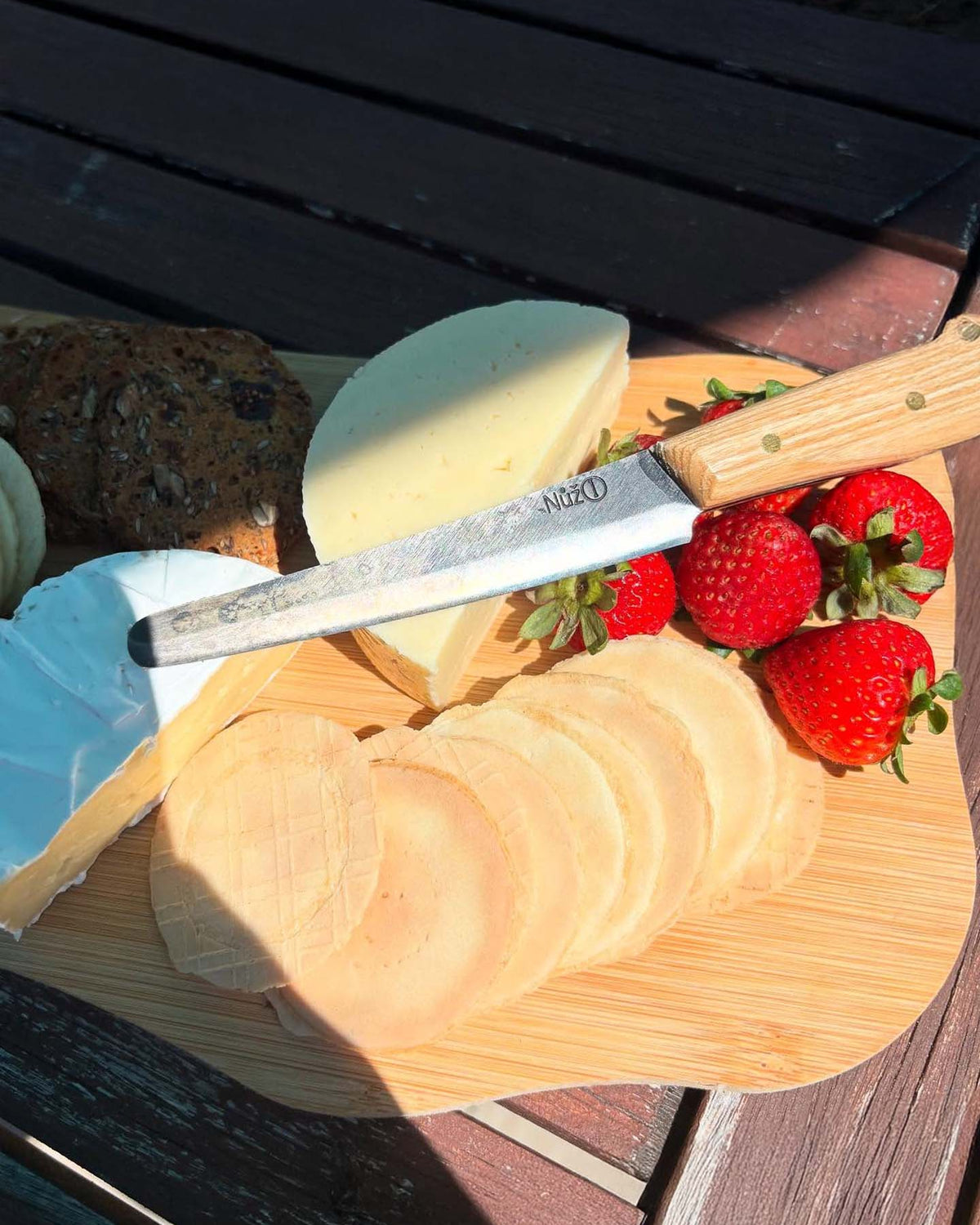The №1 – Spread Knife rests on a wooden board with assorted cheeses, round crackers, and fresh strawberries, set atop an outdoor table.